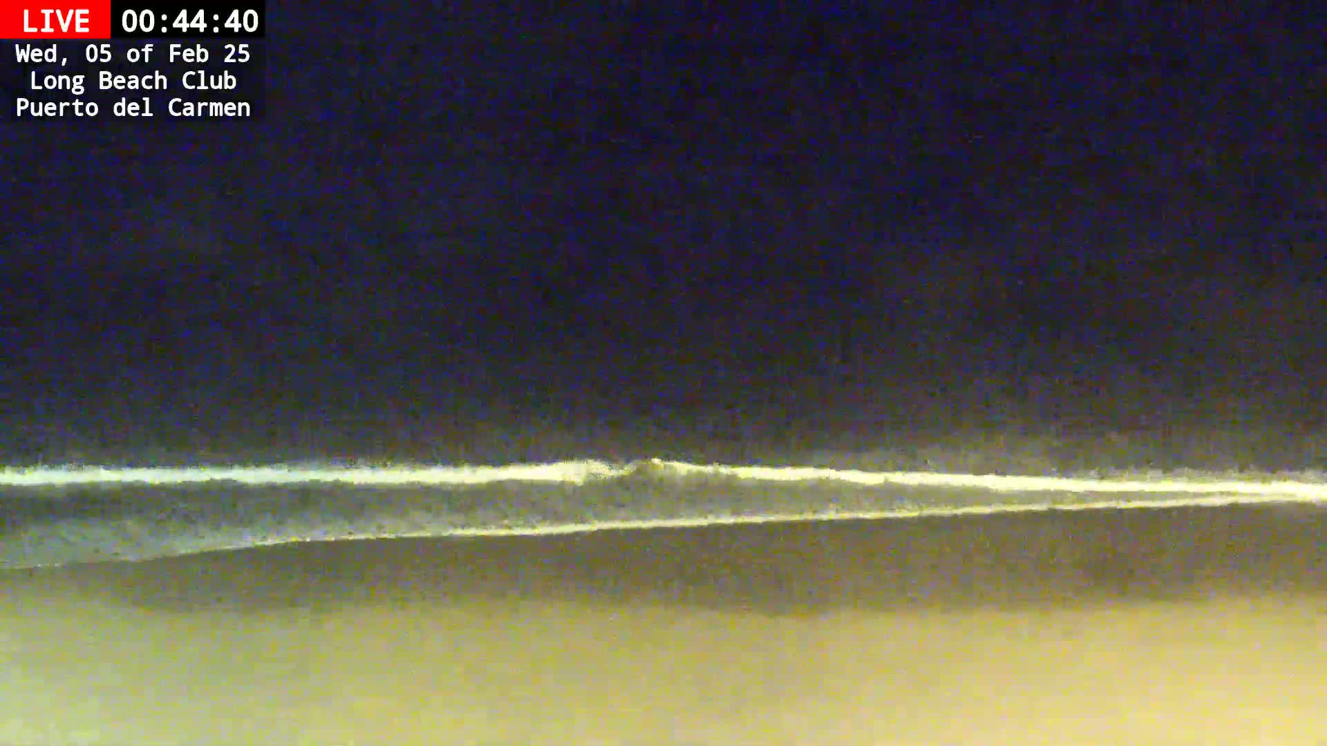 A dark, nighttime beach scene shows a thin line of white waves breaking on the shore under a dark sky.