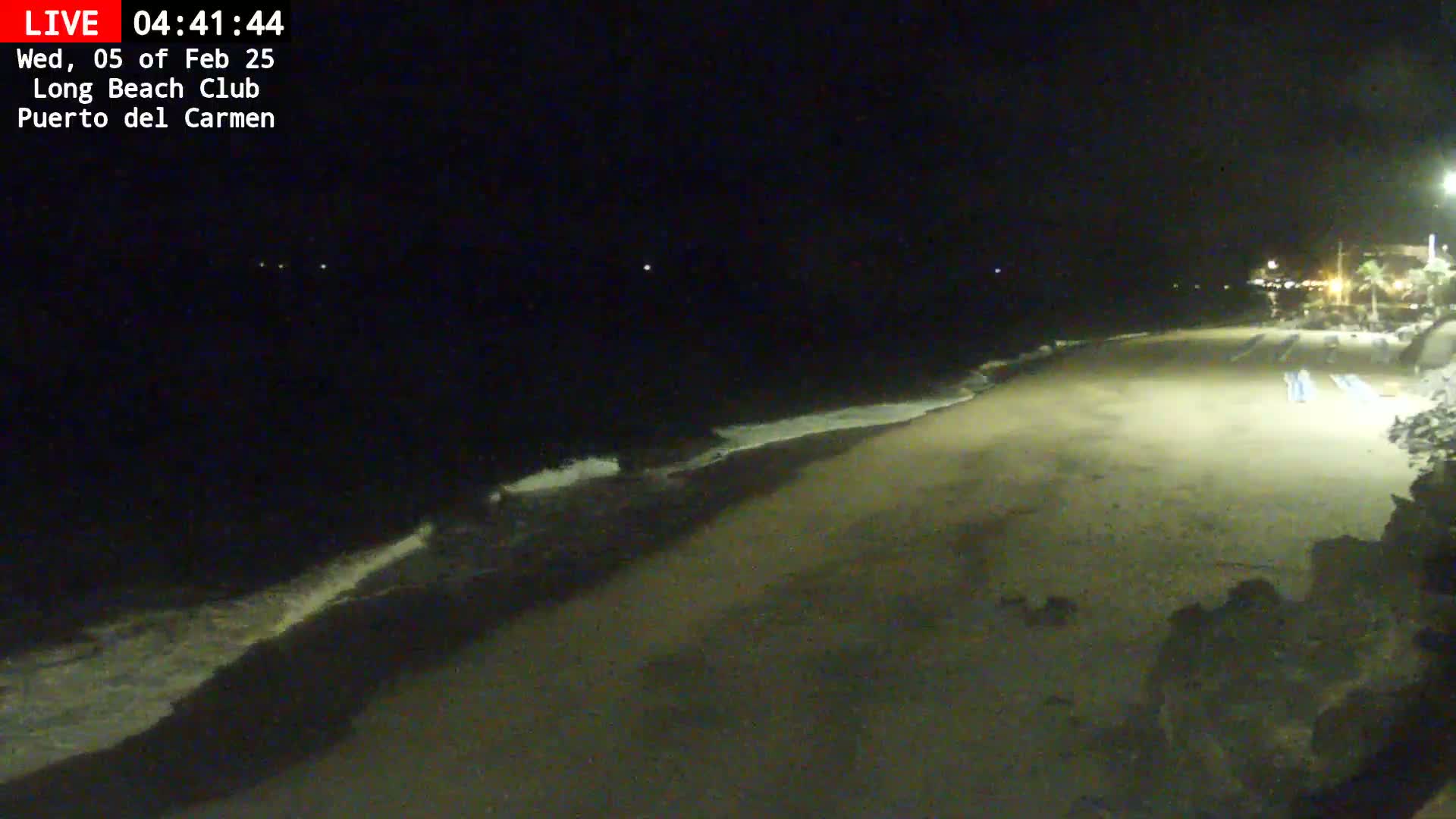 A dark, sandy beach at night with gentle waves lapping the shore, illuminated by artificial light.