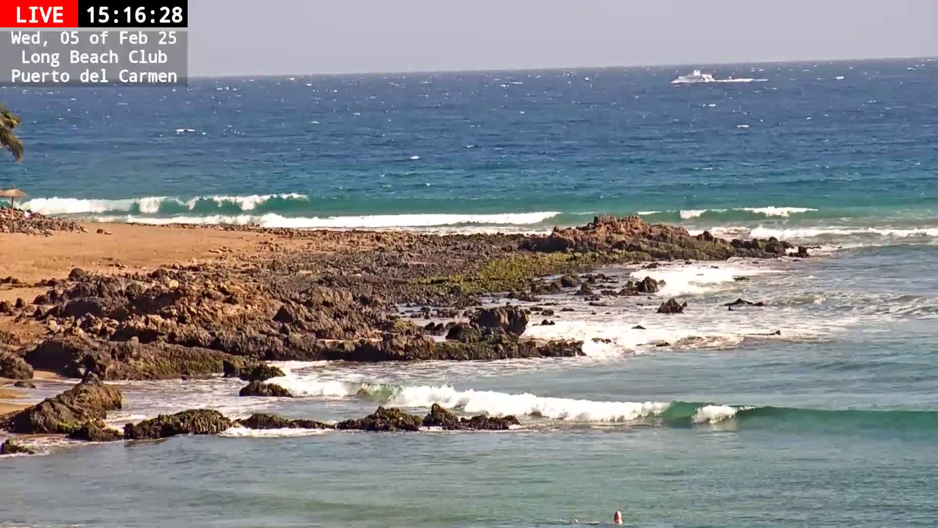 A rocky shoreline with small waves gently lapping against it, under a clear sunny sky, and a boat visible in the distance.