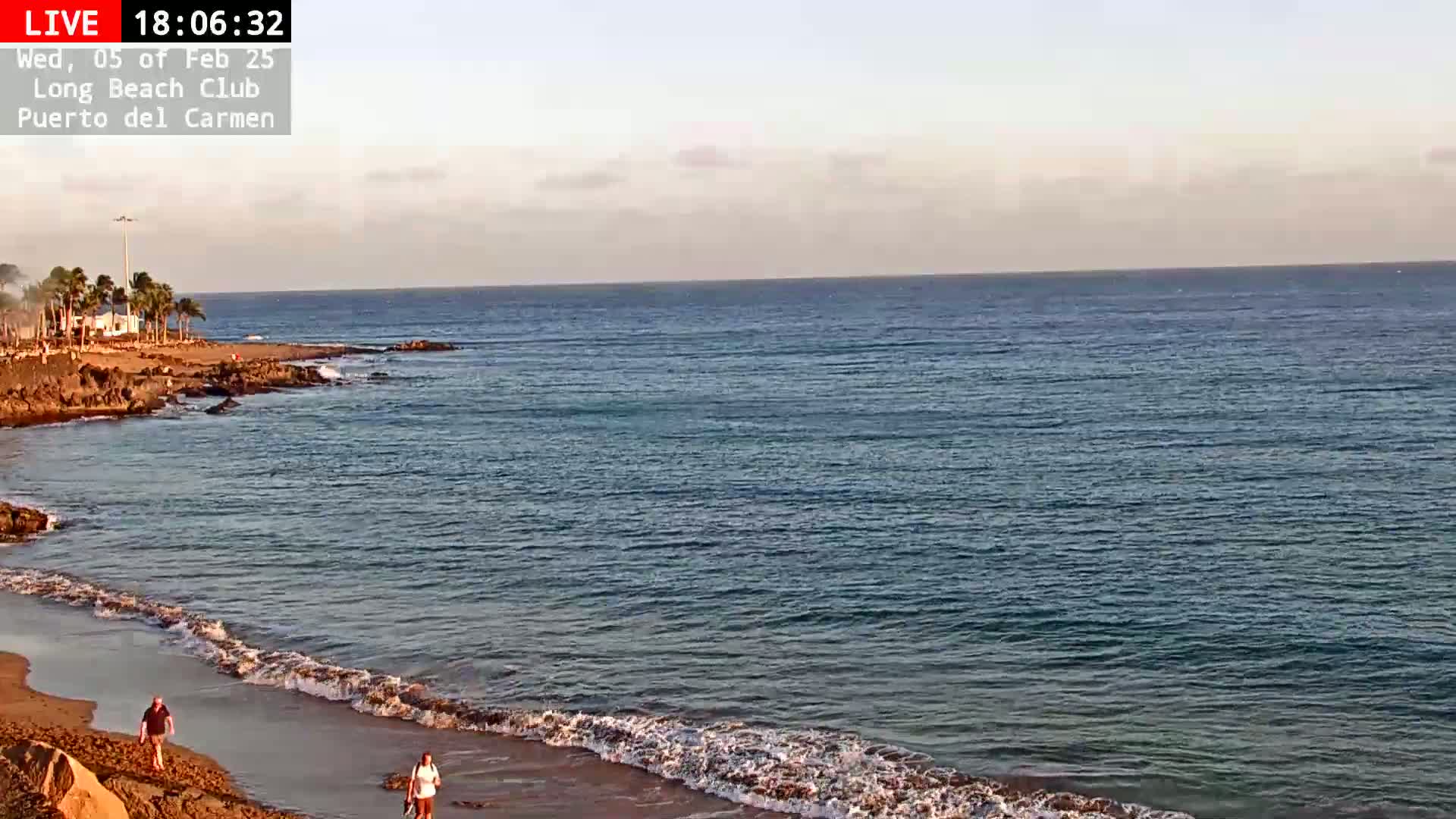 Two people walk along a sandy beach with gentle waves lapping the shore under a mostly clear sky.