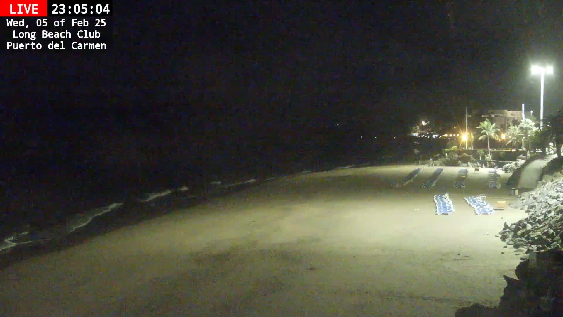 A nighttime view of a mostly empty sandy beach with rows of lounge chairs, a dark ocean with small waves, and a dimly lit building in the background under a clear, dark sky.