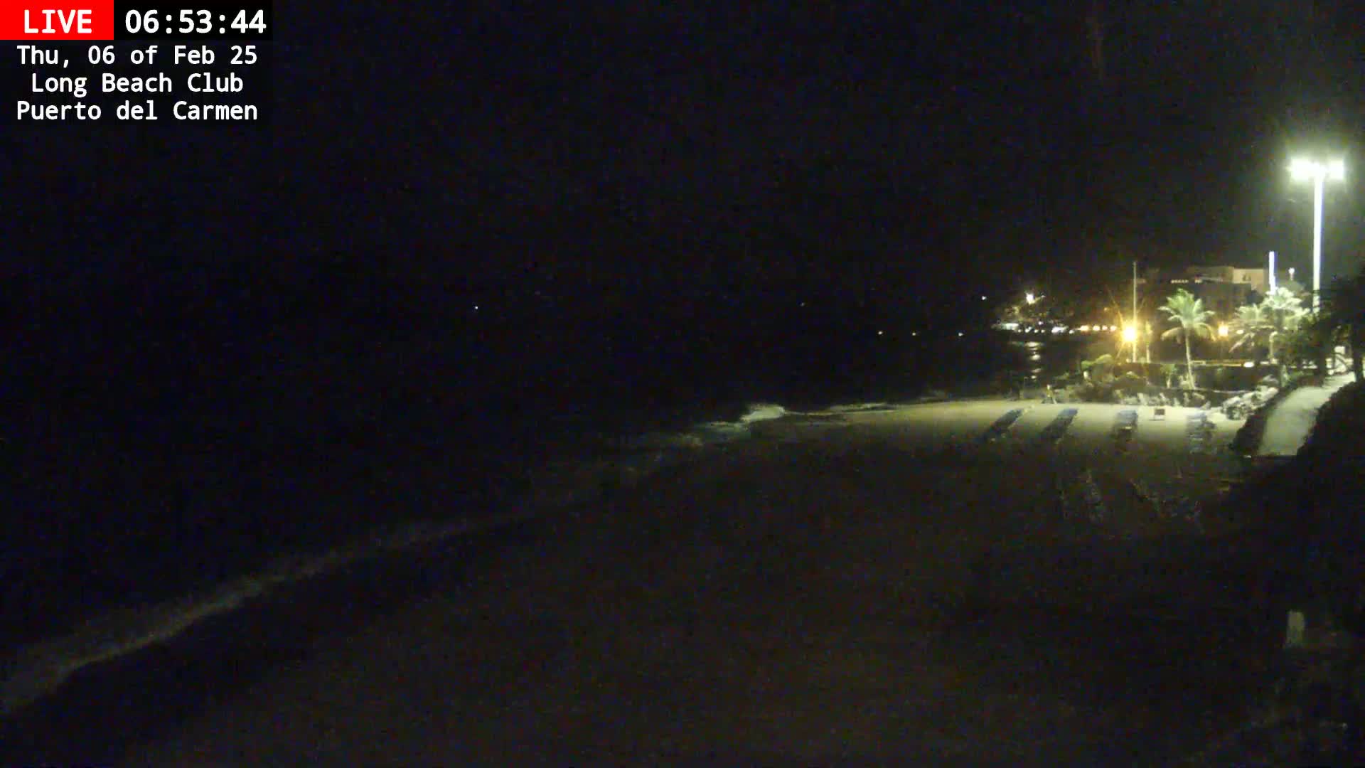 A dark, nighttime beach scene shows a mostly empty sandy shoreline with a few lounge chairs under artificial lights, and a dark ocean with small waves.