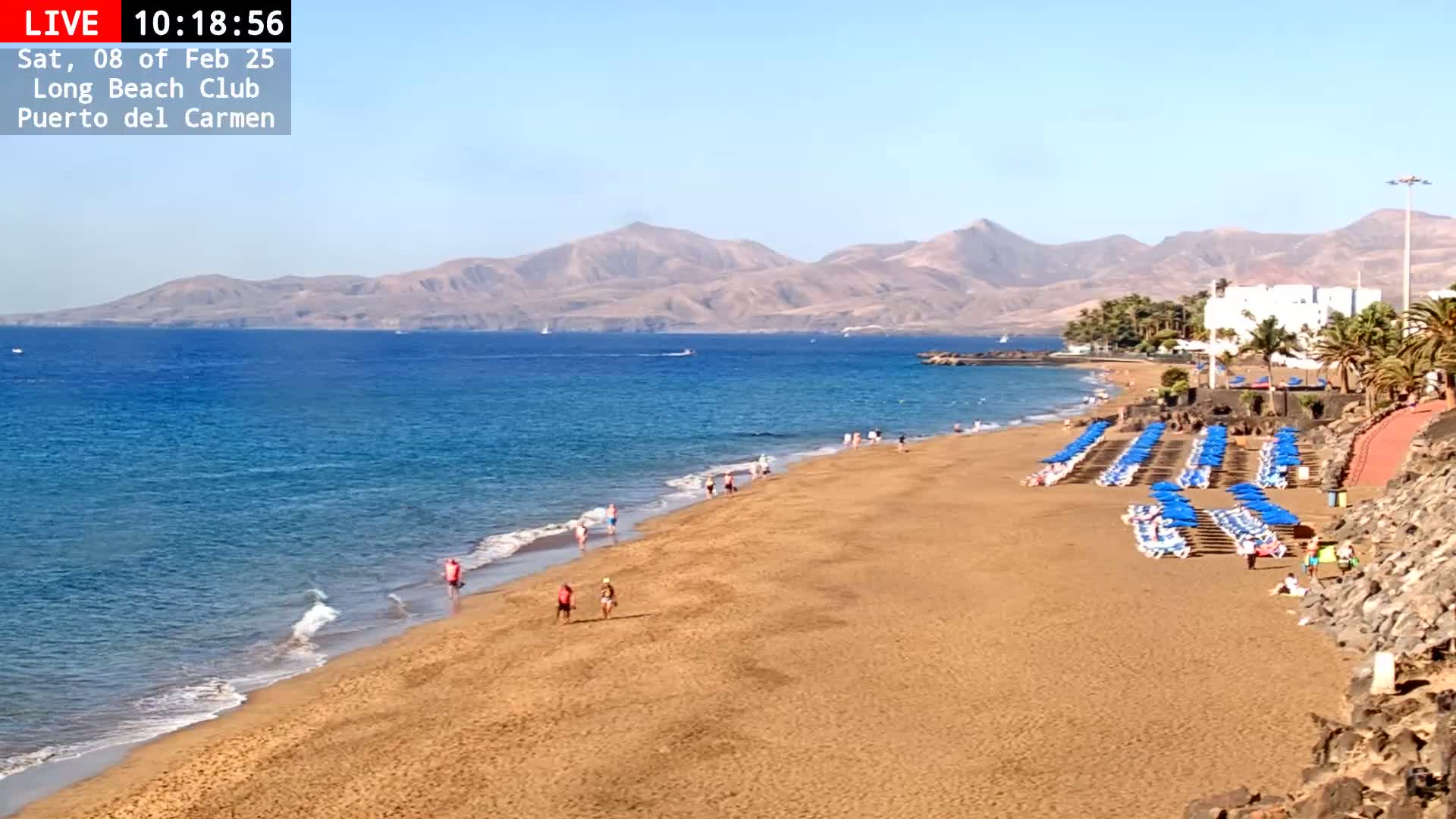 A sandy beach with blue umbrellas and a few people on it stretches toward a calm, blue ocean and brown mountains under a clear, sunny sky.
