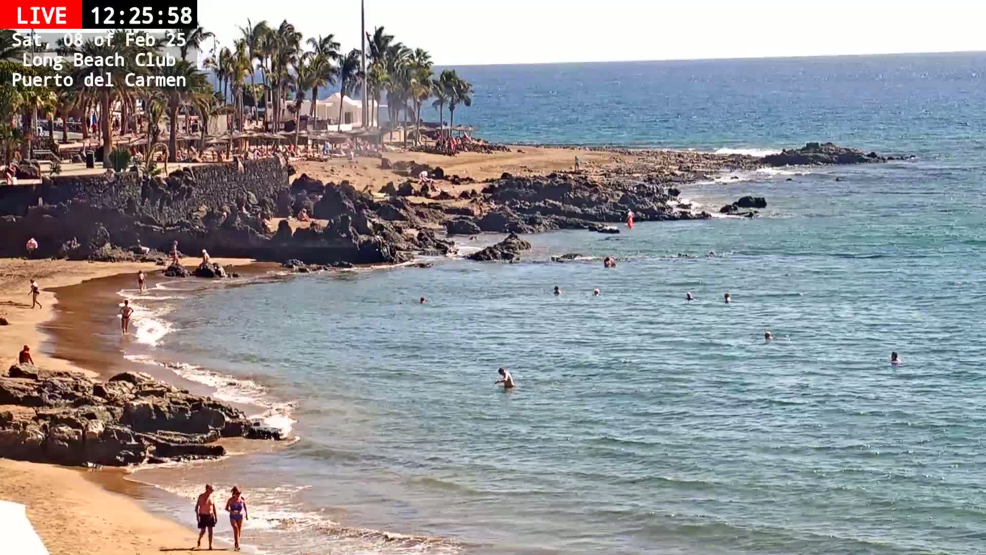 A sandy beach with dark volcanic rocks and calm, clear blue water is populated by a few swimmers and sunbathers on a sunny day.