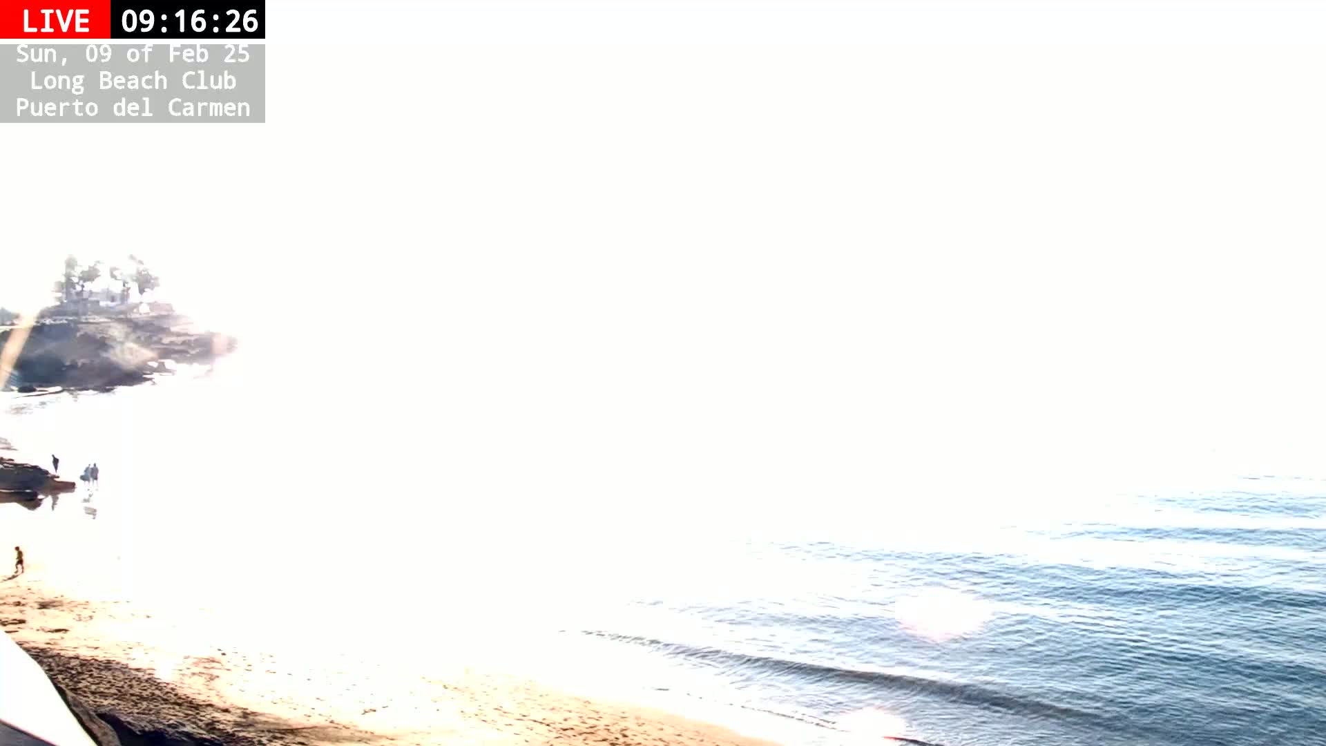 A bright, sunny beach scene shows a sandy shore, calm ocean waves, and a few people near some rocks.