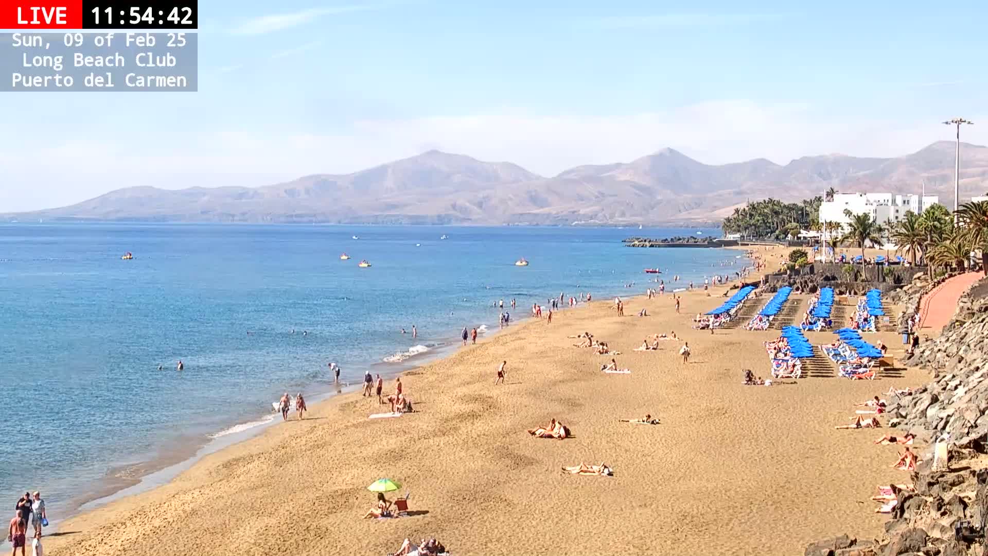 A sandy beach with many people sunbathing and swimming in a calm ocean under a clear, sunny sky, backed by mountains.
