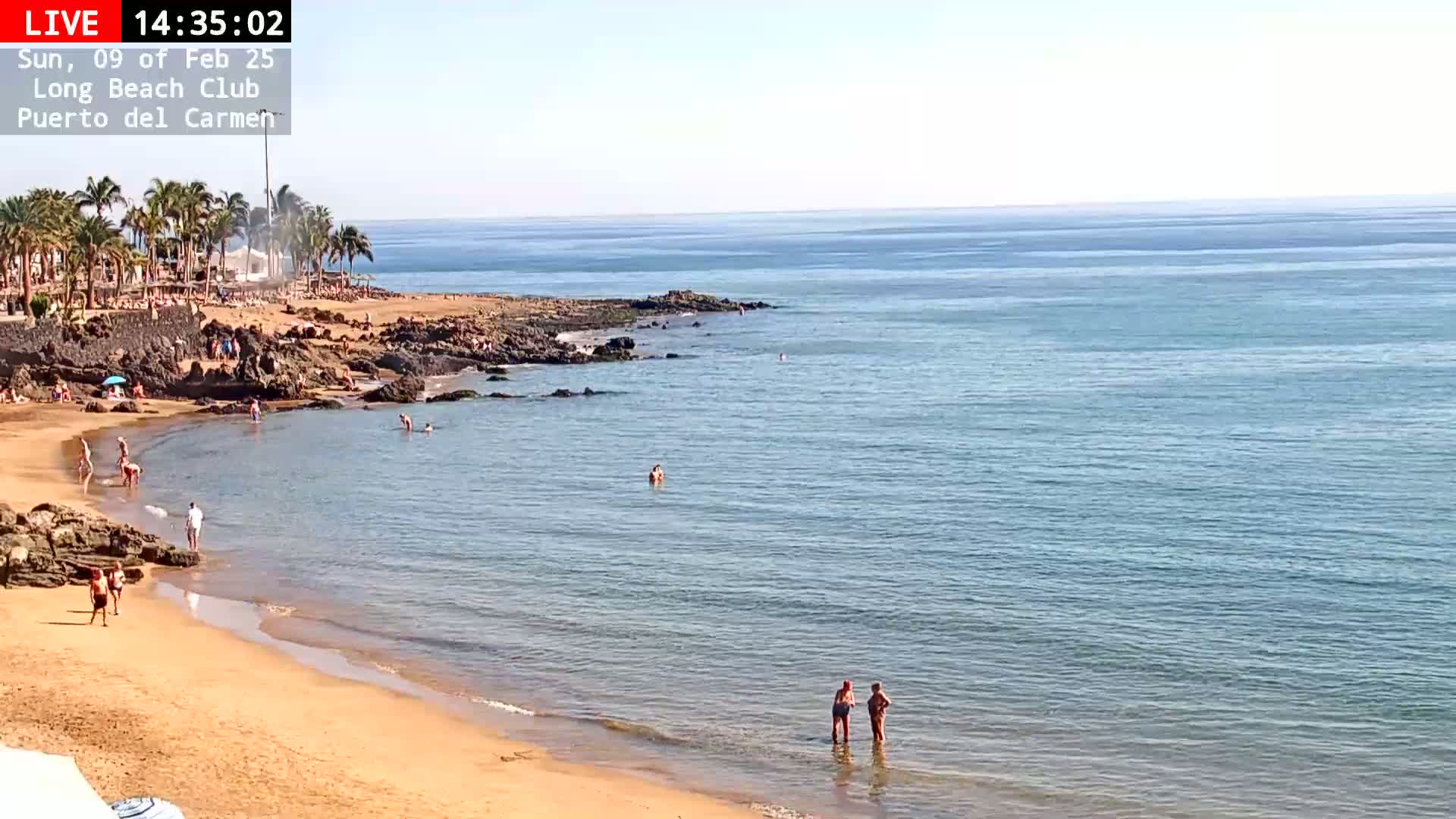 A sandy beach with calm, clear blue water, dotted with people swimming and sunbathing, under a sunny sky.