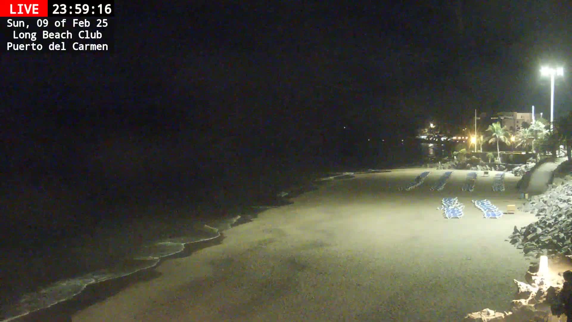 A nighttime beach scene shows gently lapping waves on a sandy shore with several rows of empty lounge chairs under artificial lights.