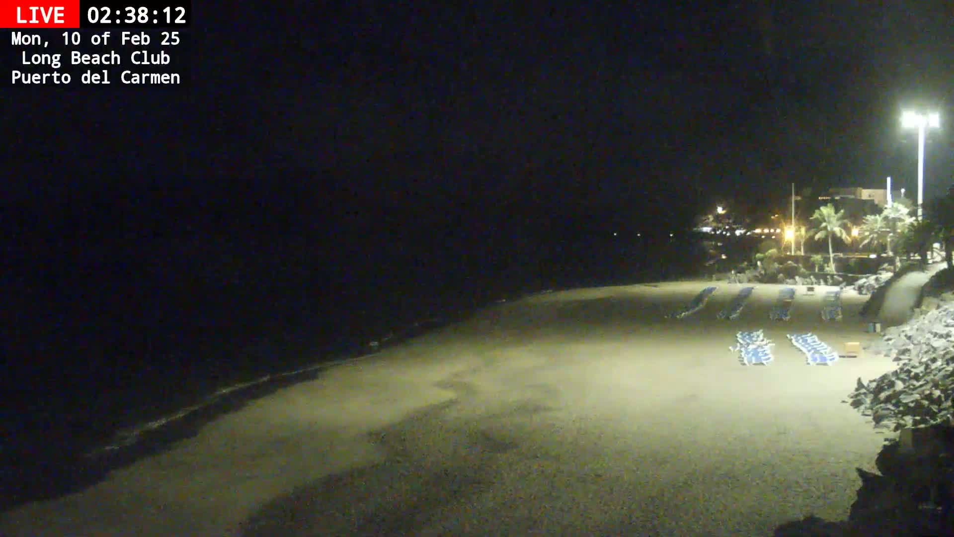 A mostly empty, sandy beach at night is dimly lit, with a few rows of lounge chairs and a calm ocean visible in the background.