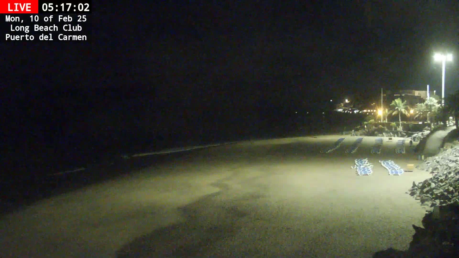 A dark, nighttime beach scene shows a sandy shore with several empty lounge chairs near a rocky embankment under a mostly clear sky.