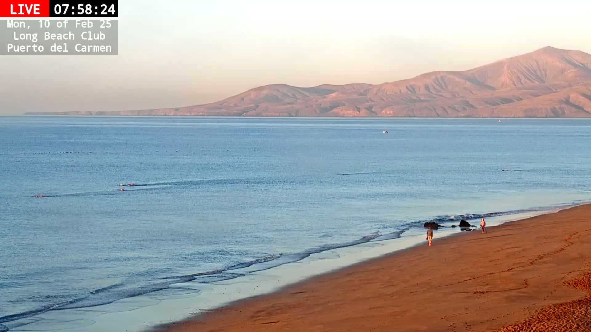 A sandy beach with a calm ocean and a few people walking along the shore, under a clear, light-pink sky with distant mountains visible.