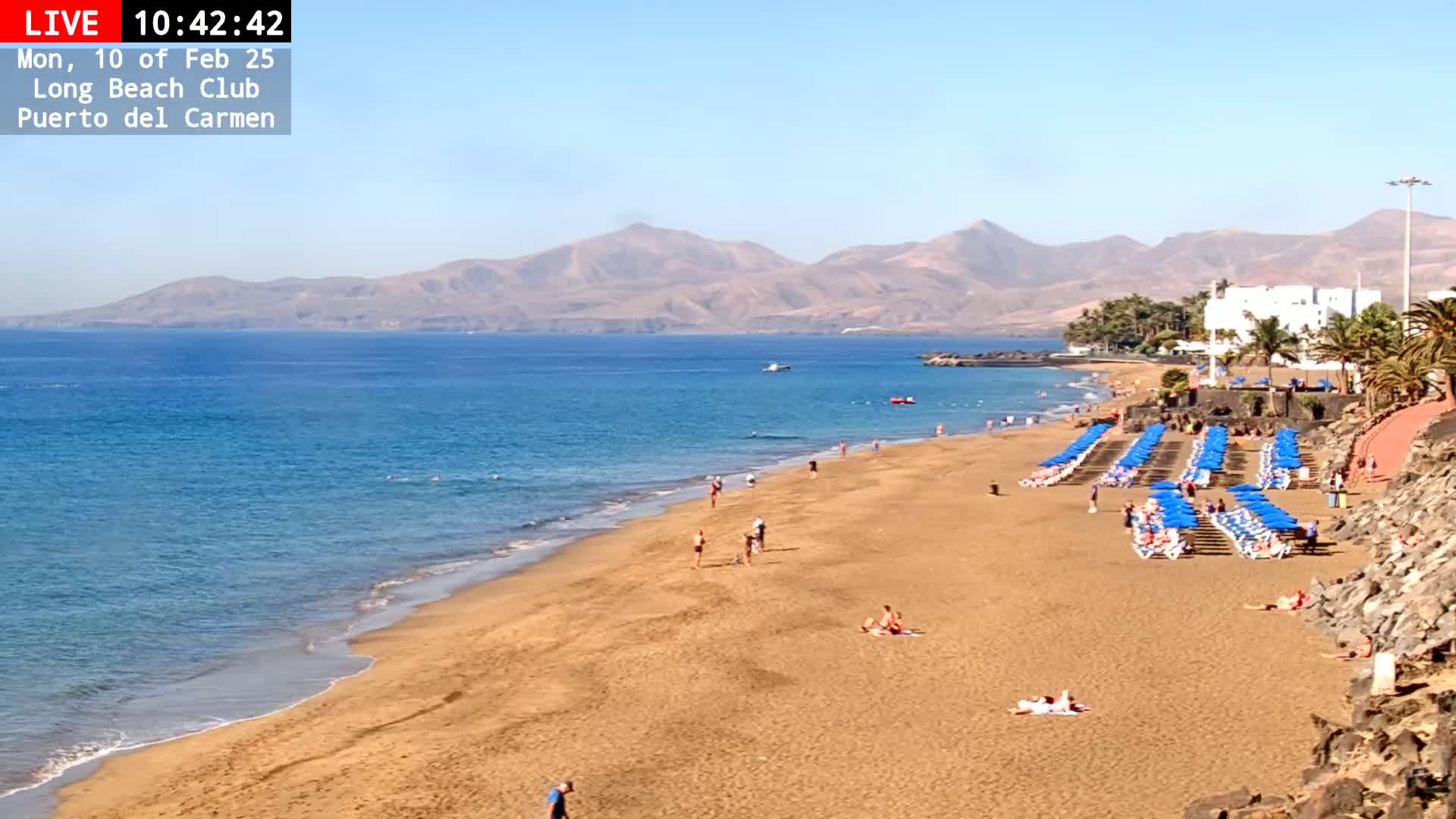 A sandy beach with blue umbrellas and lounge chairs stretches along a calm, blue ocean under a clear sky, backed by mountains.