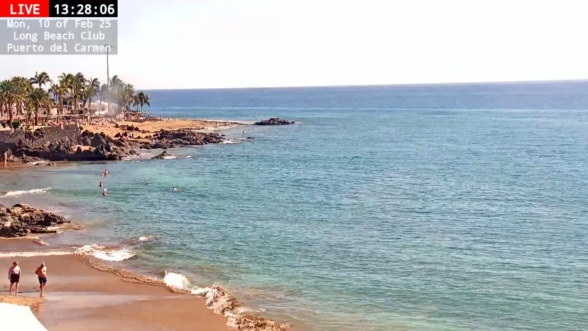 A sandy beach with calm, clear water and several people swimming and walking near a rocky shoreline on a sunny day.