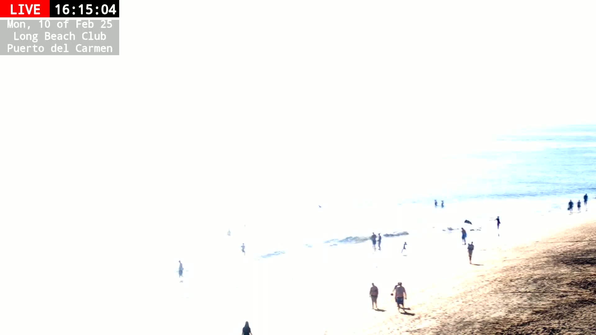 A sunny beach scene shows people walking along the shore and in the shallow water.