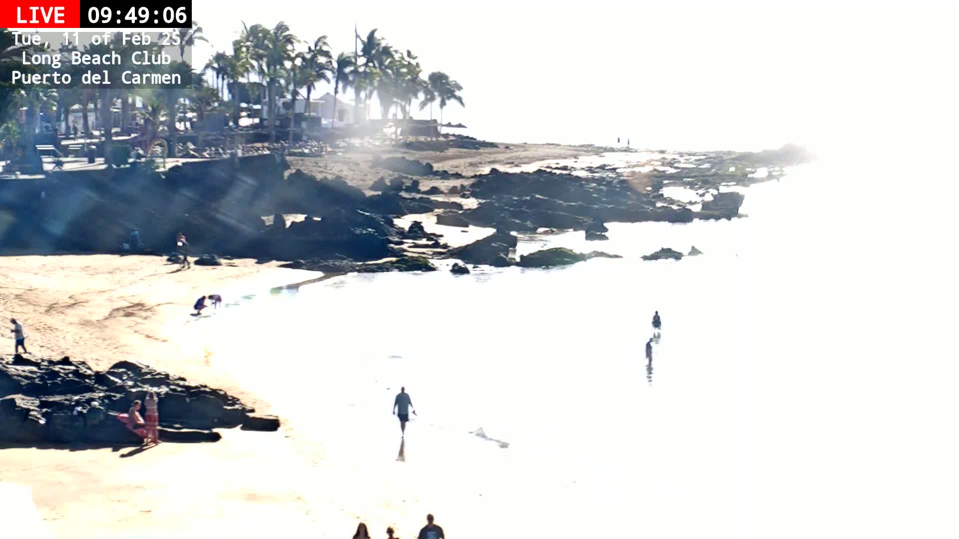A sandy beach with dark rocks and shallow, calm water is sparsely populated with people on a bright, sunny day.