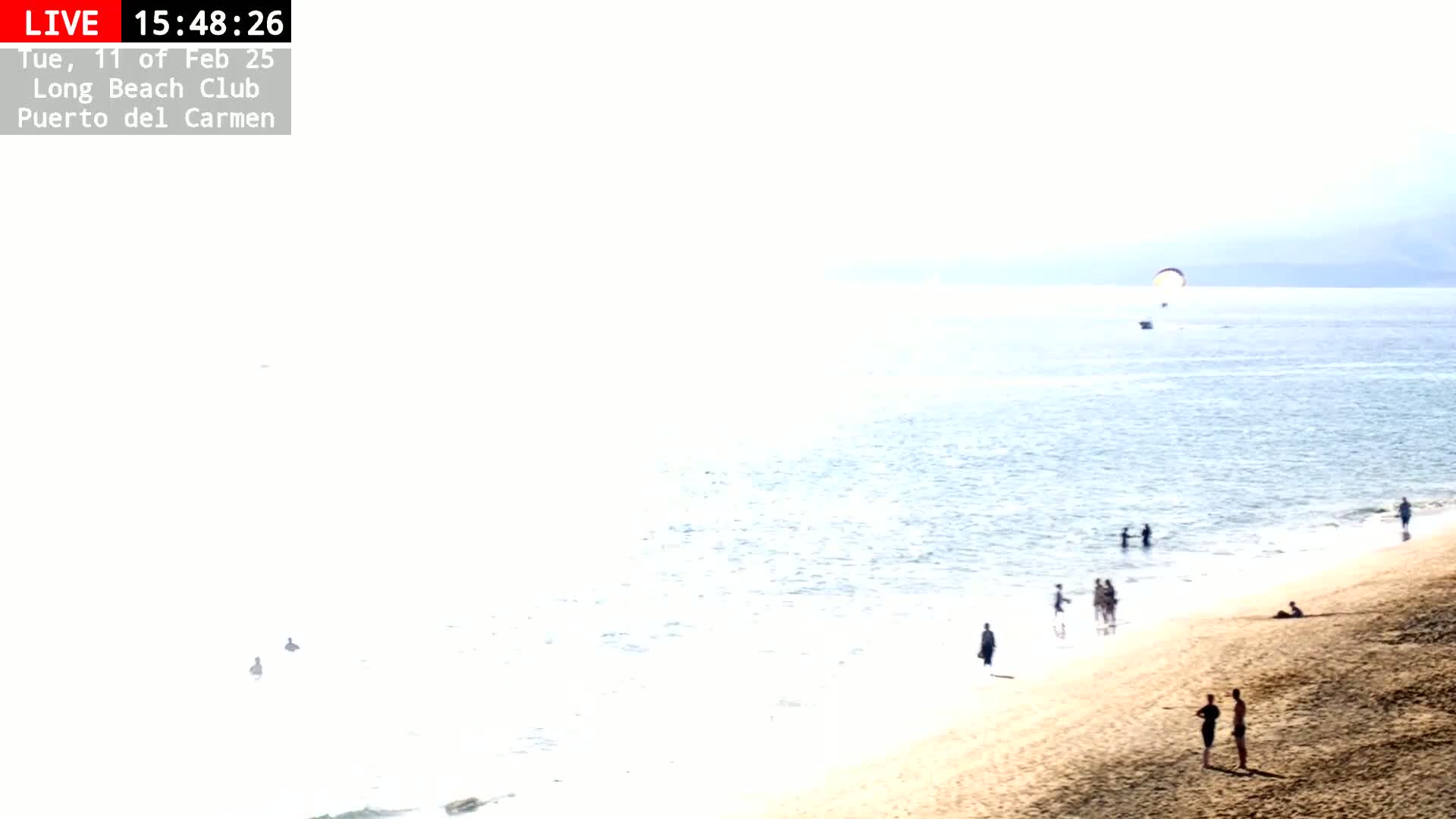 A sandy beach with a few people on it and in the water, under a bright, sunny sky, with a parasail visible in the distance.