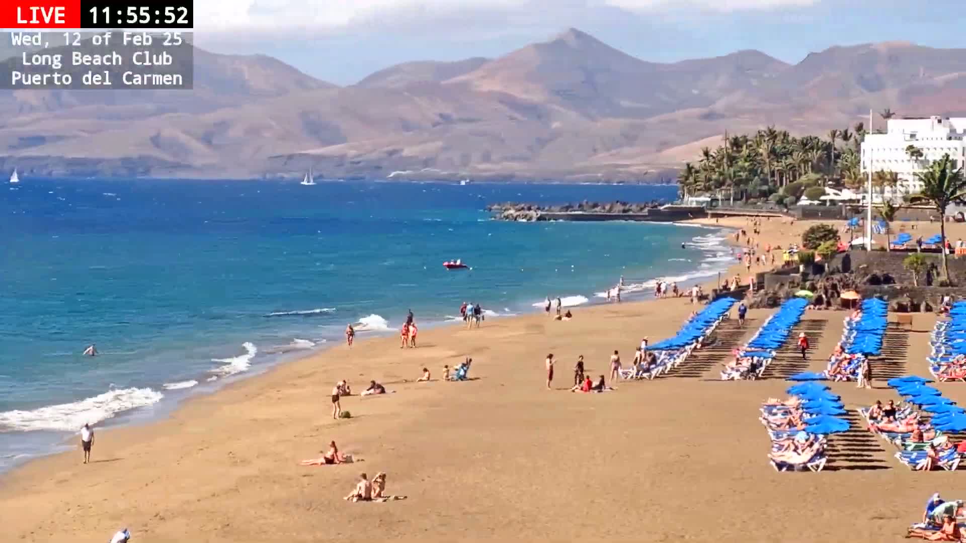 A sandy beach with many people sunbathing and swimming, lined with blue umbrellas and chairs, extends to a calm, blue ocean under a mostly sunny sky, backed by mountains.