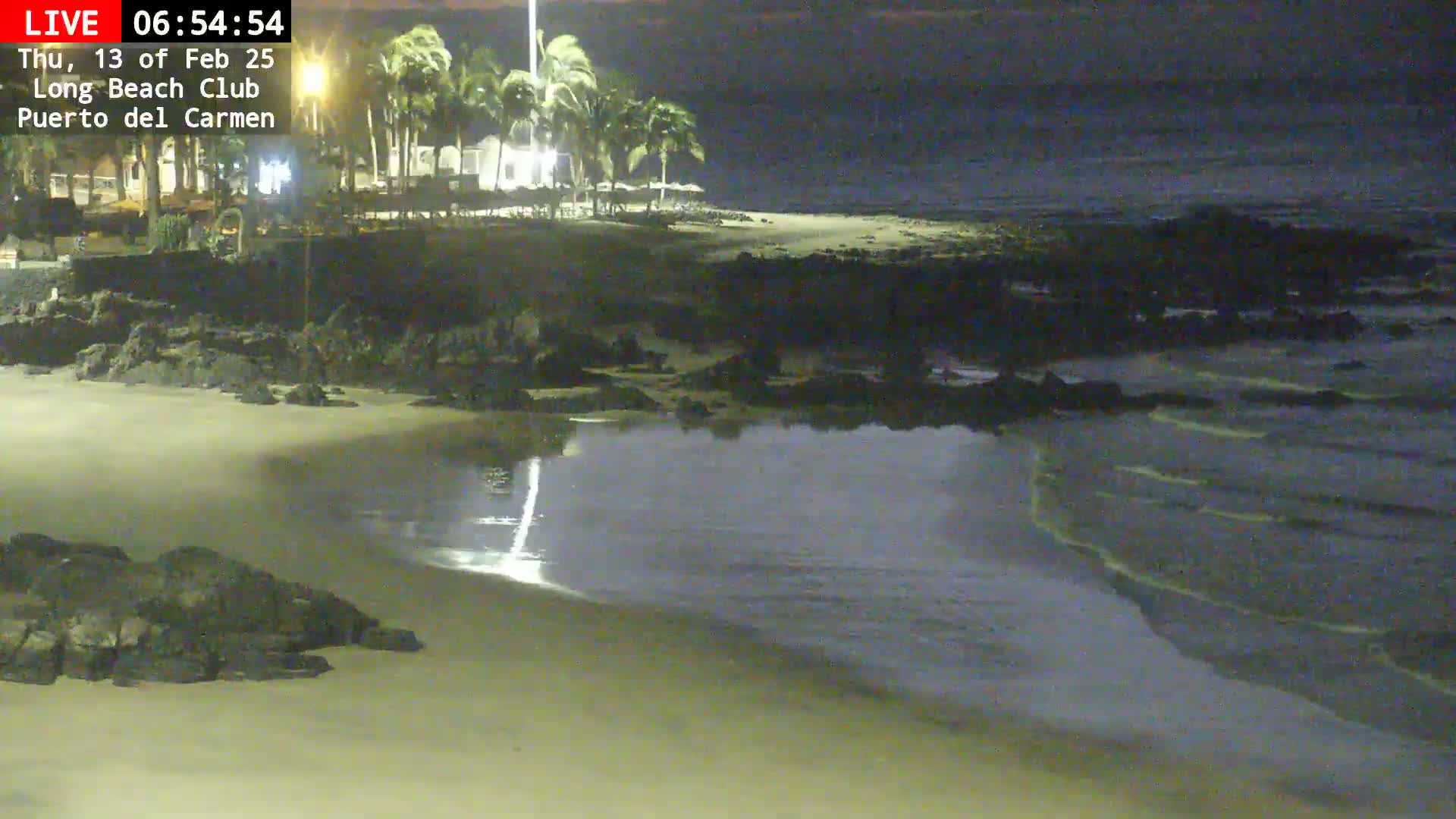A sandy beach at night with gentle waves lapping the shore, palm trees and buildings visible in the background under a dark sky.