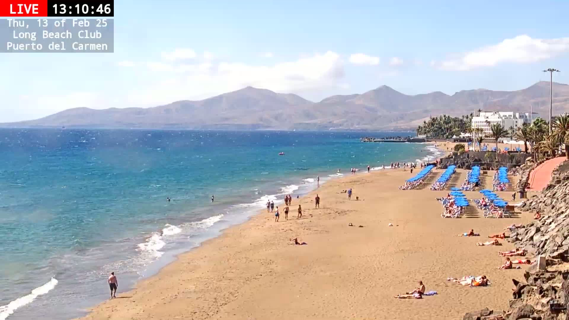 A sandy beach with blue umbrellas and sunbathers stretches along a turquoise ocean, backed by mountains under a partly cloudy sky.