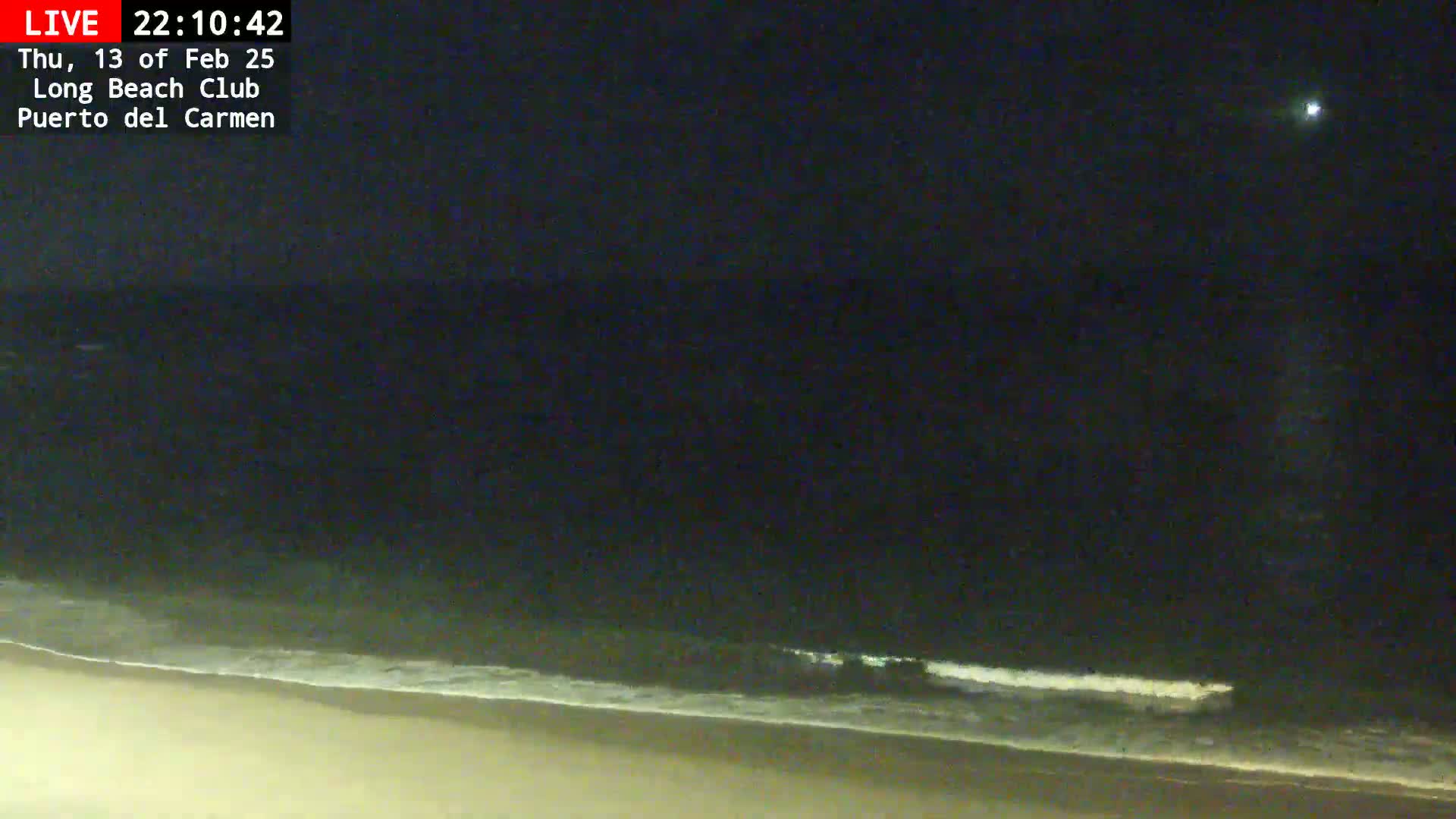 A nighttime view shows a dark ocean with small waves lapping a sandy beach under a clear, dark sky.