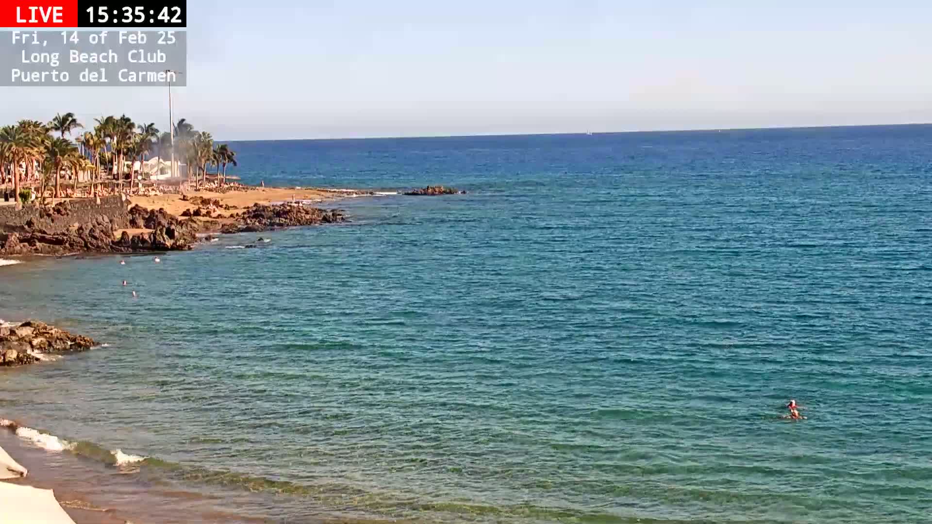 A sunny day at a beach with clear, teal water shows a sandy shore lined with palm trees and dark rocks, with a few people swimming or wading in the ocean.
