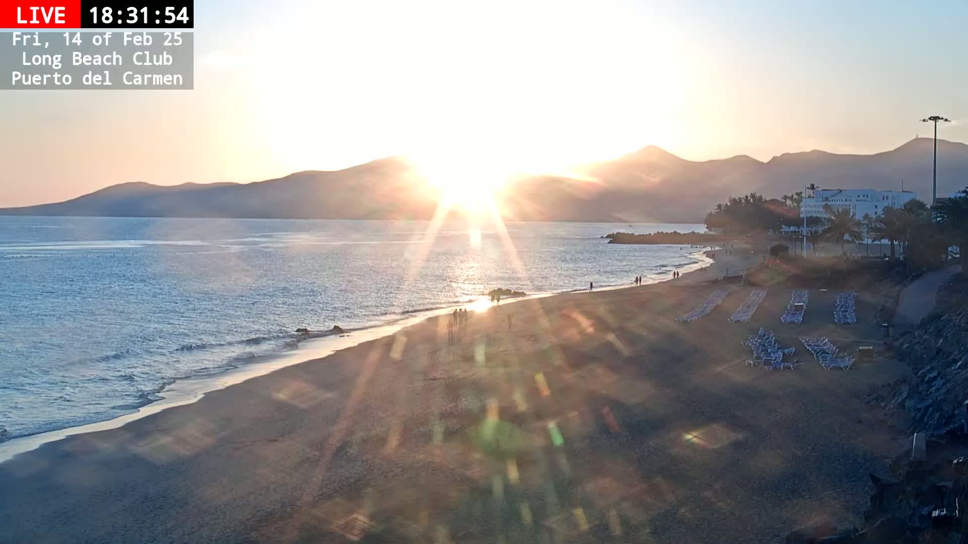 A sandy beach with a few people walking along the shore at sunset, with mountains visible in the distance.