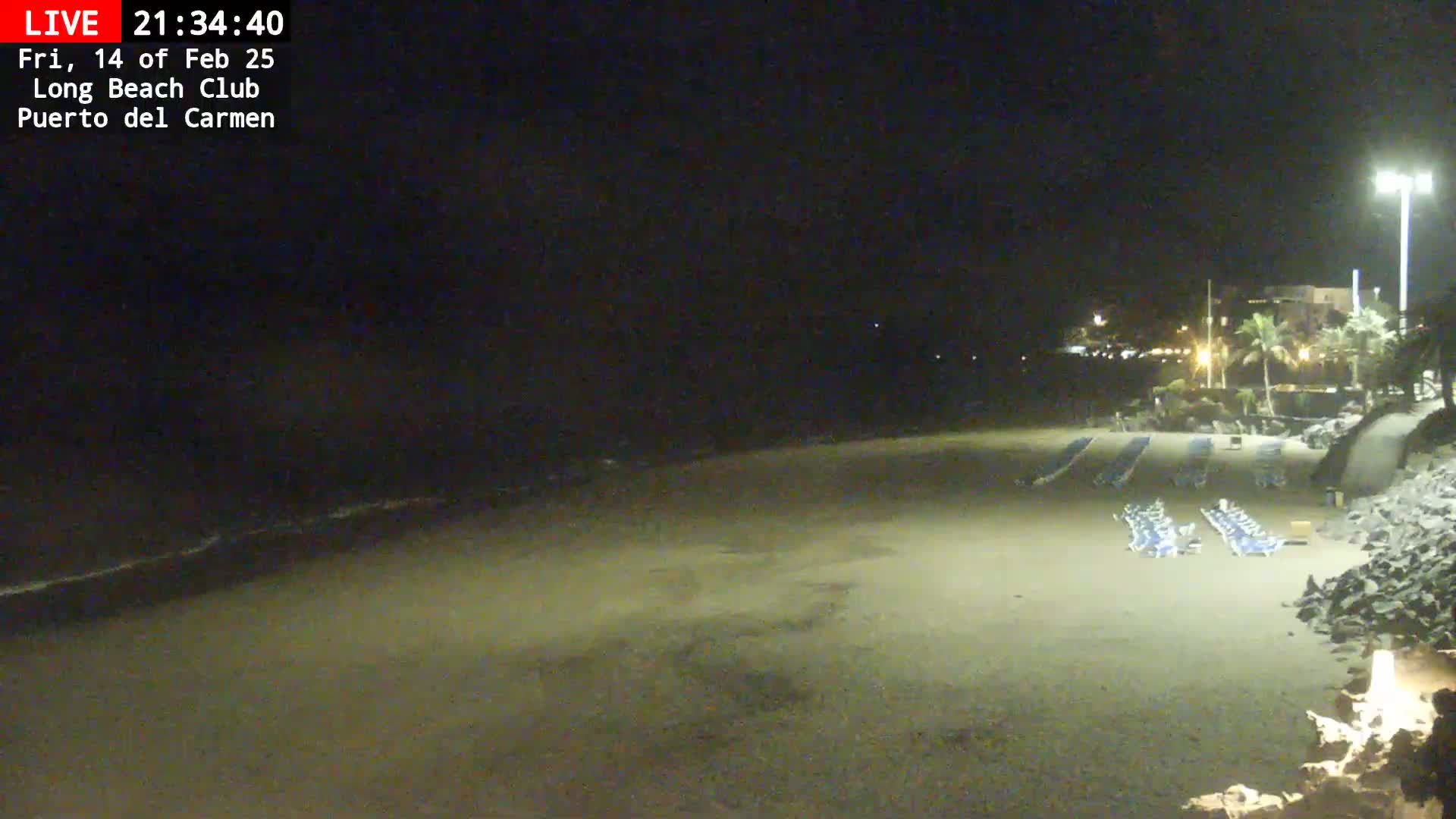 A dark, sandy beach at night is mostly empty except for several rows of folded beach chairs near the shore under a mostly clear sky.