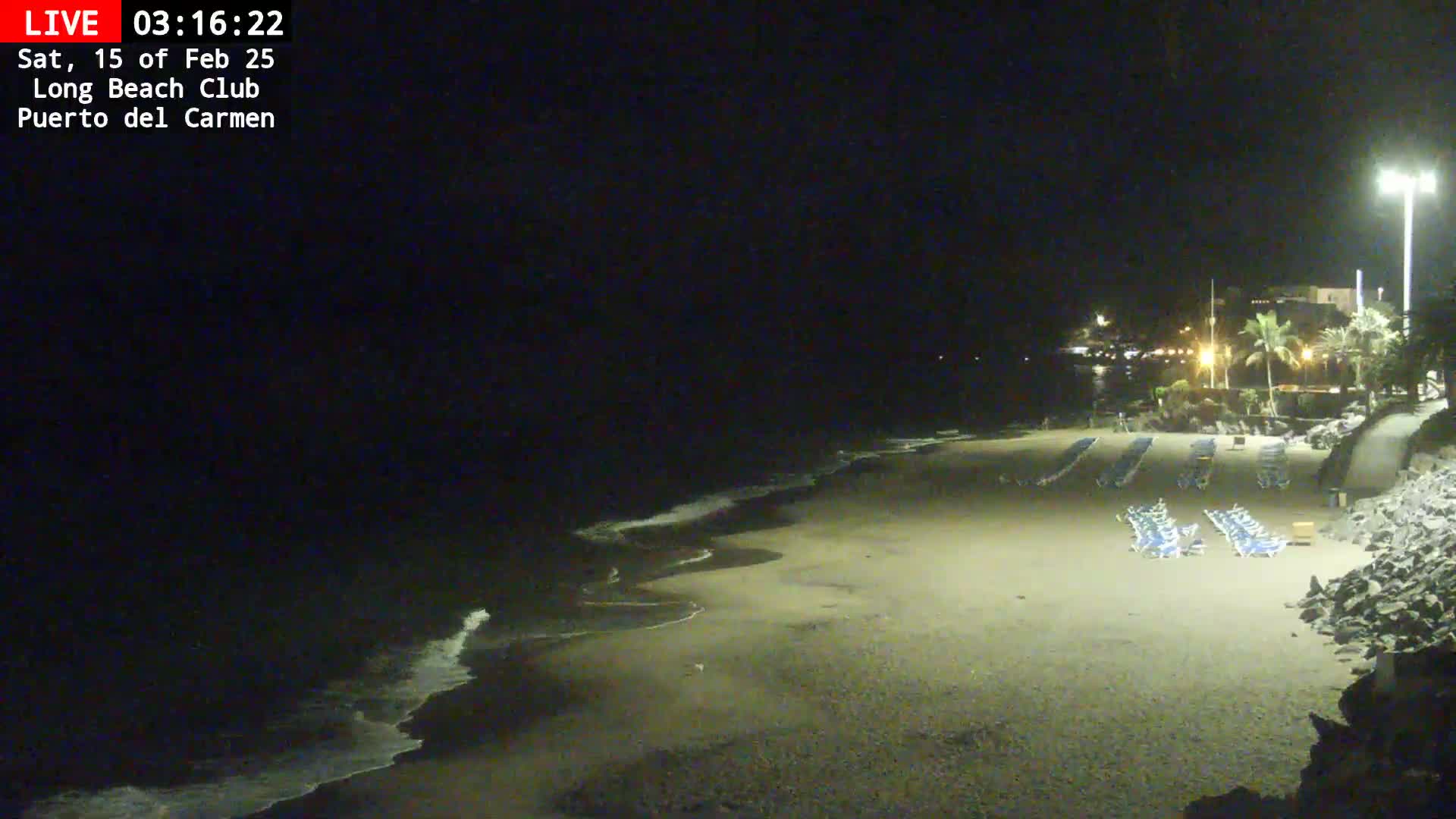 A dark, nighttime beach scene shows gently lapping waves on a sandy shore with several rows of empty lounge chairs under artificial light.