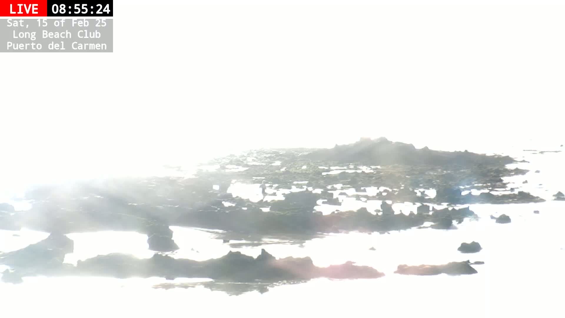 A bright, hazy sun illuminates dark rocks and shallow water at the shoreline.