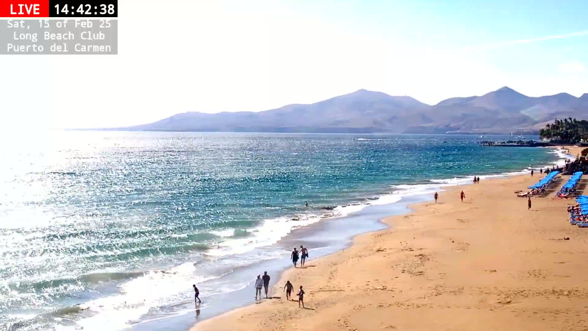 A sandy beach with people walking along the shore and rows of blue beach umbrellas, with a wavy ocean and mountains in the background under a sunny sky.