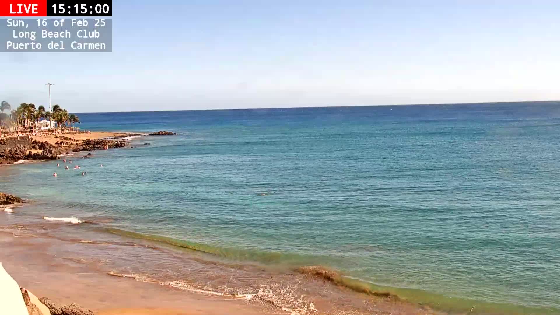 A sandy beach with clear, turquoise water and several people swimming under a sunny sky.