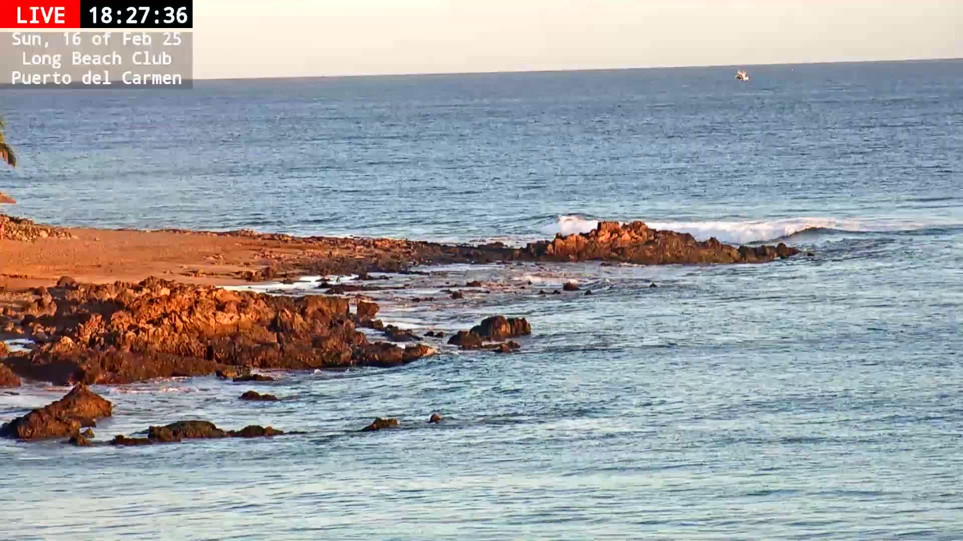 Dark-colored rocks jut out from a calm, blue ocean under a clear sky.