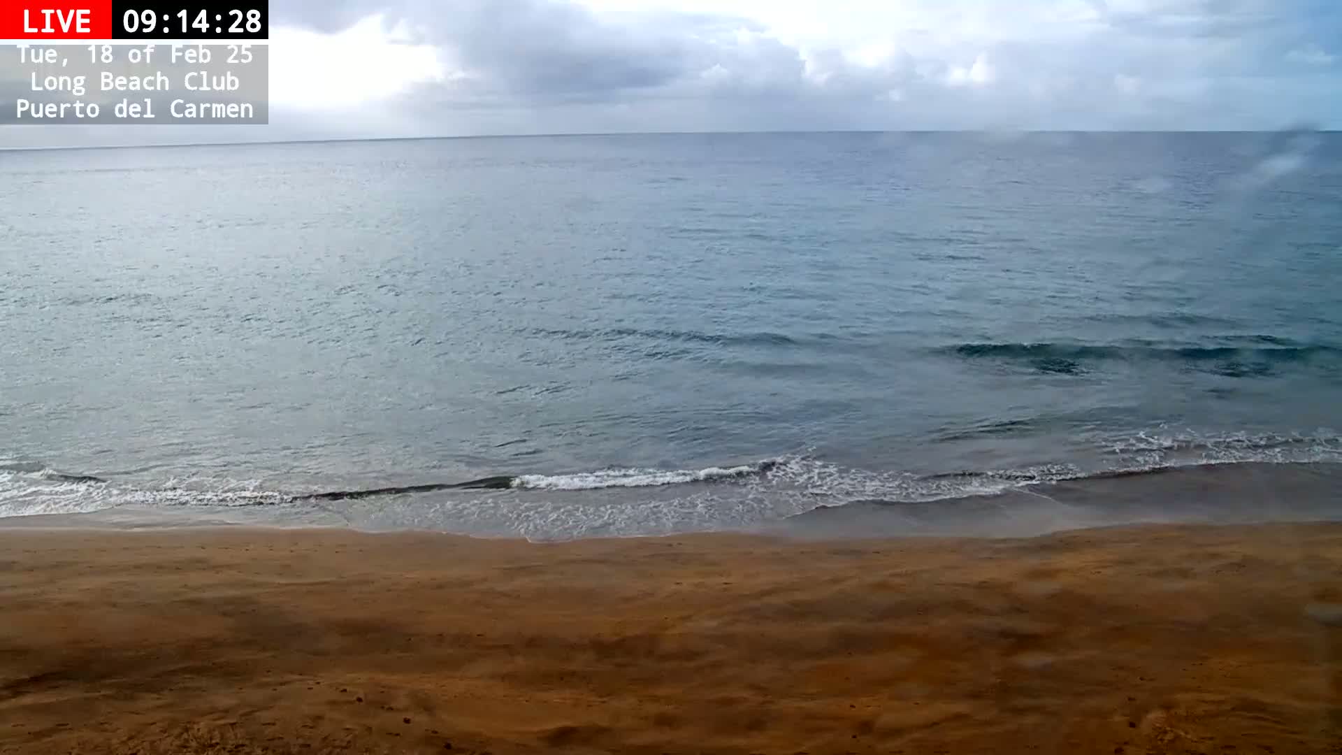 A sandy beach meets a calm, gray-blue ocean under a mostly cloudy sky.