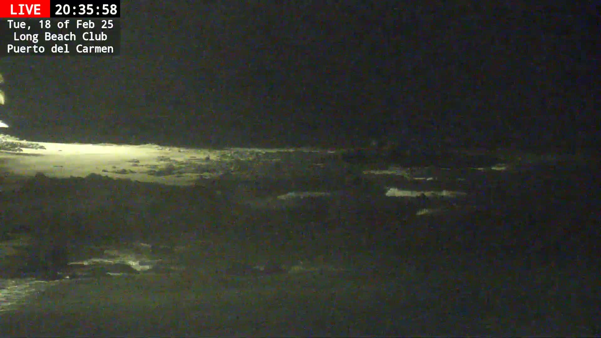 A dark, nighttime beach scene shows a dimly lit shoreline with gentle waves lapping against the shore.