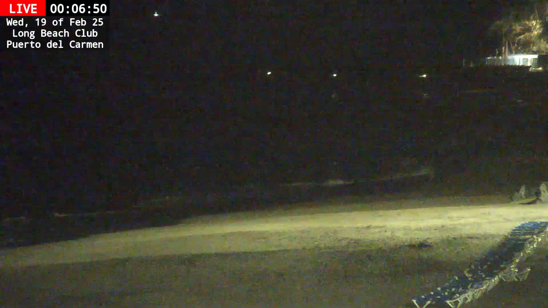A dark, nighttime beach scene shows a sandy shore with several lounge chairs in the lower right corner, under a clear, dark sky.