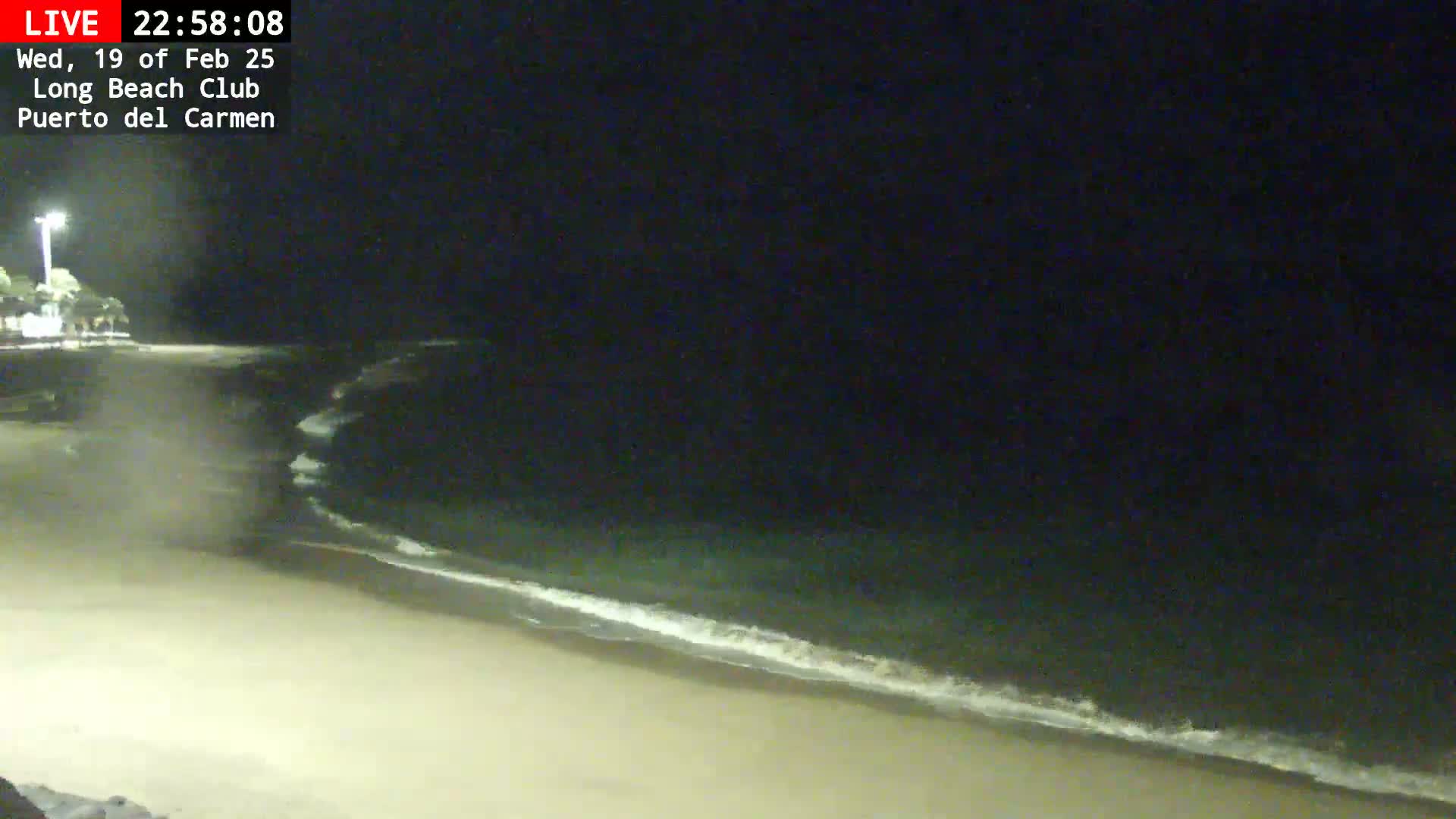 A dark, nighttime beach scene shows gently lapping waves on a sandy shore under a clear, starless sky.