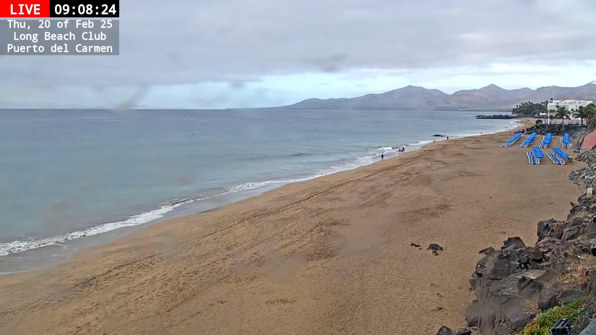 A long sandy beach with blue lounge chairs and a calm ocean under an overcast sky is seen, with mountains visible in the distance.
