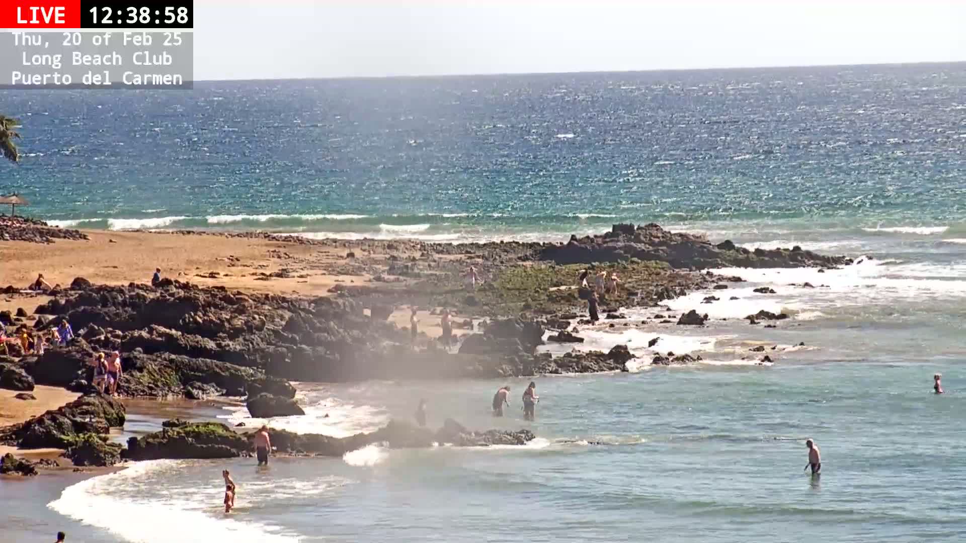 A sunny day at a rocky beach with calm waves and several people swimming and wading in the shallow water.