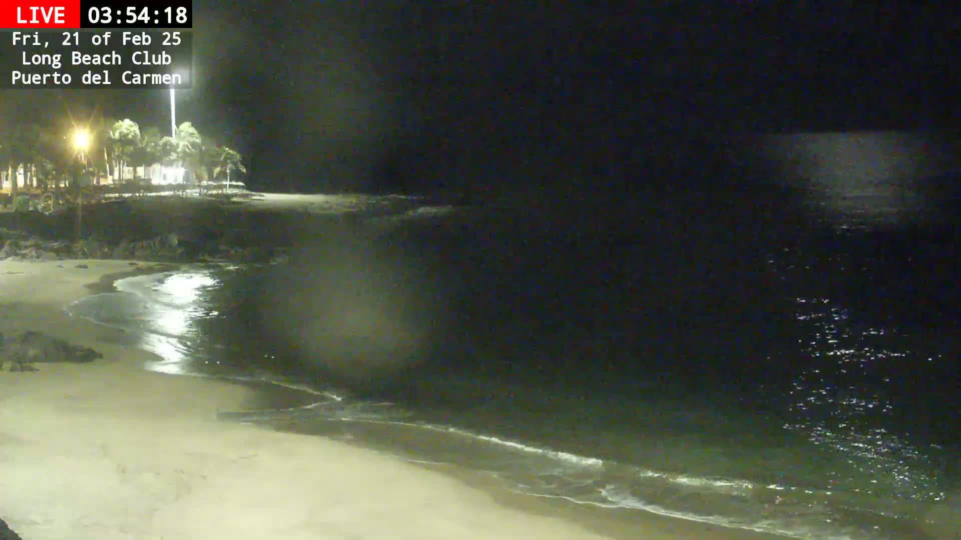 A nighttime view of a mostly empty sandy beach with calm waves gently lapping the shore under a dark sky.