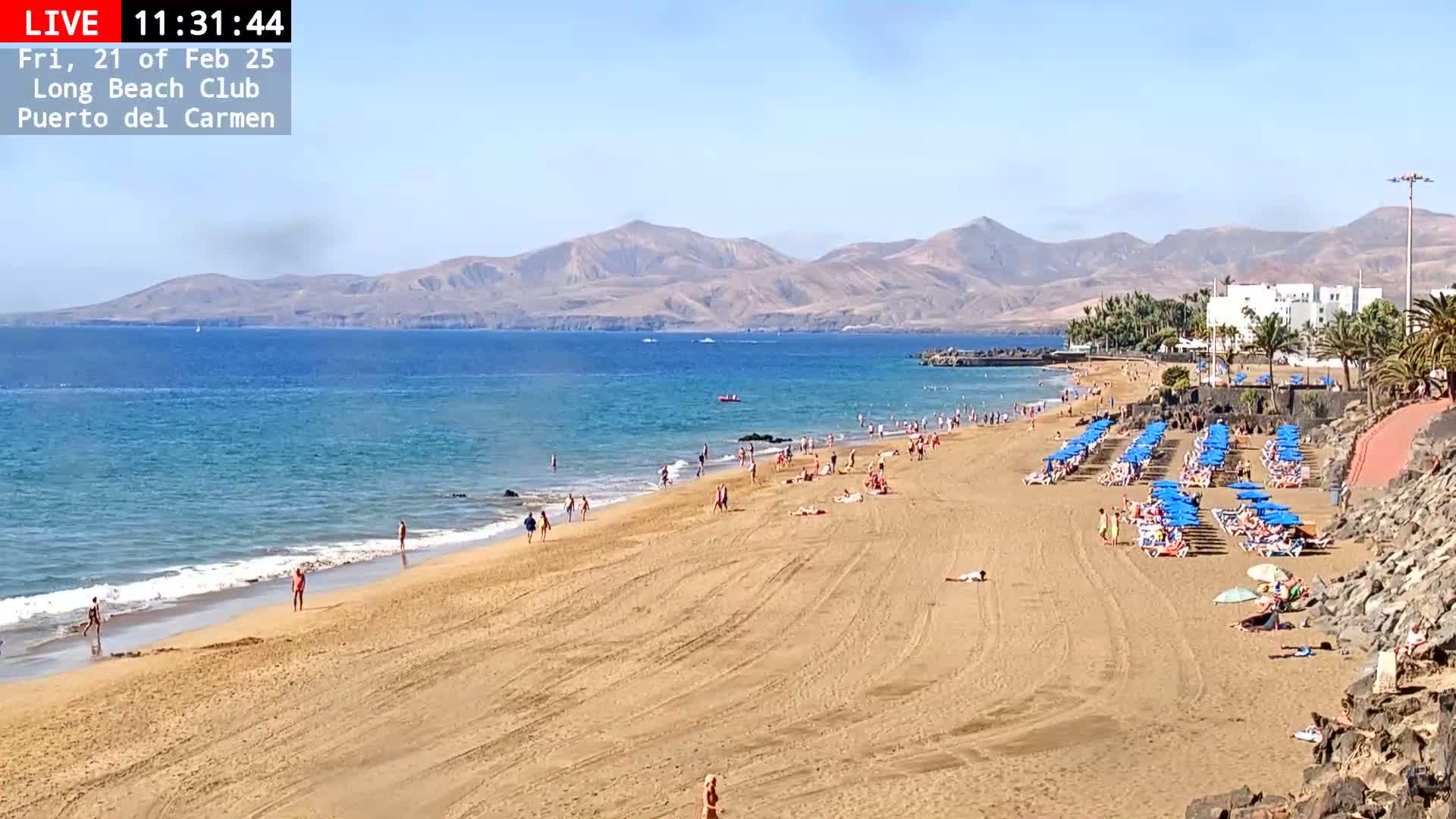 A sandy beach with numerous people sunbathing and swimming in a calm ocean under a clear, sunny sky, backed by mountains.