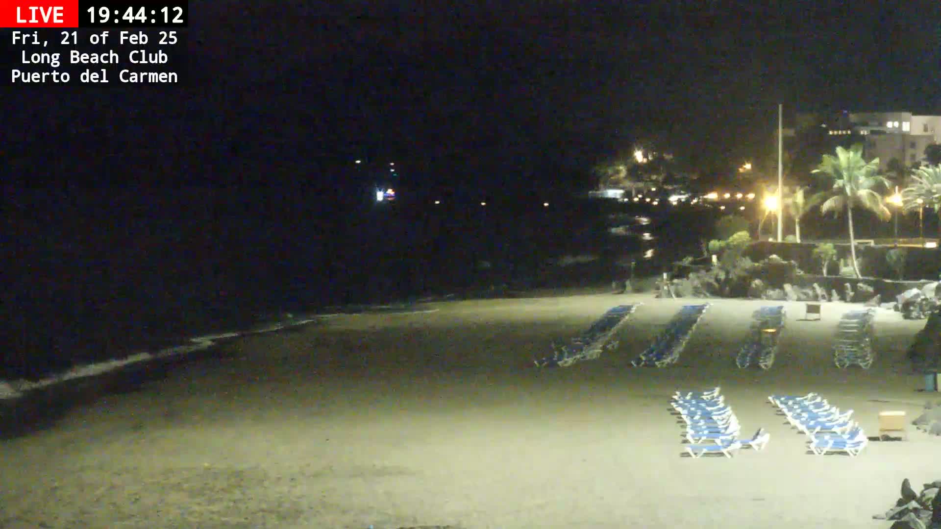 At night, an empty sandy beach with rows of lounge chairs is gently lapped by the ocean under a dark sky.