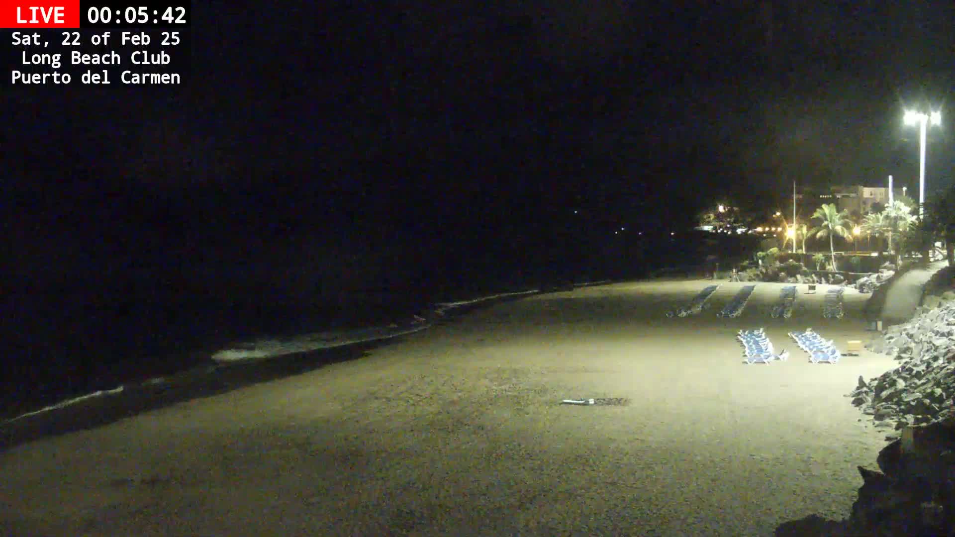 A dark, sandy beach at night with gently lapping waves, rows of empty lounge chairs, and a dimly lit building in the background.