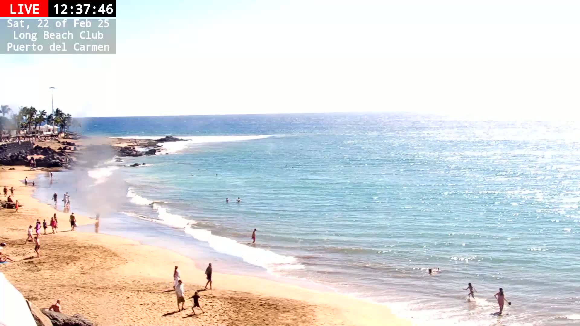 A sunny beach scene shows people relaxing and swimming in calm, clear turquoise water.