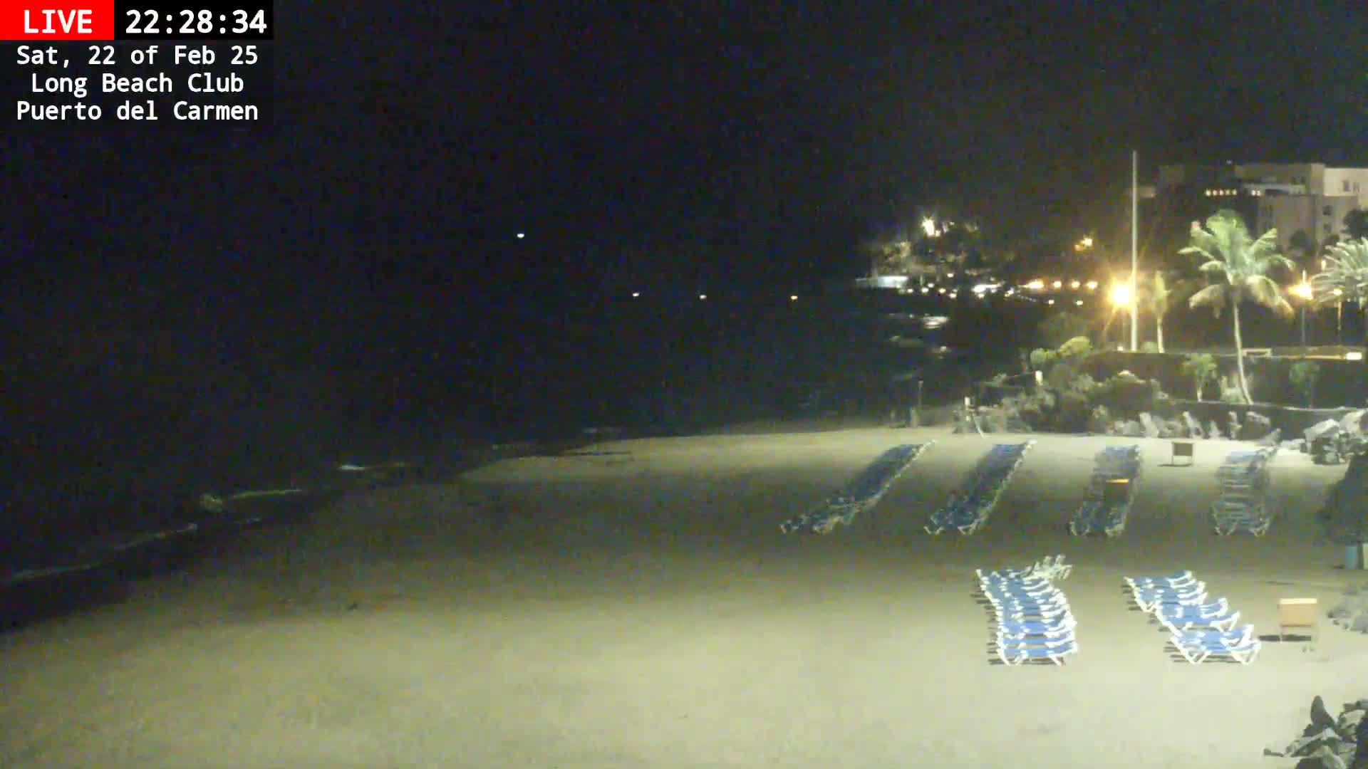 A dark sandy beach at night is mostly empty except for rows of blue lounge chairs, with a building and palm trees visible in the background under a clear, dark sky.