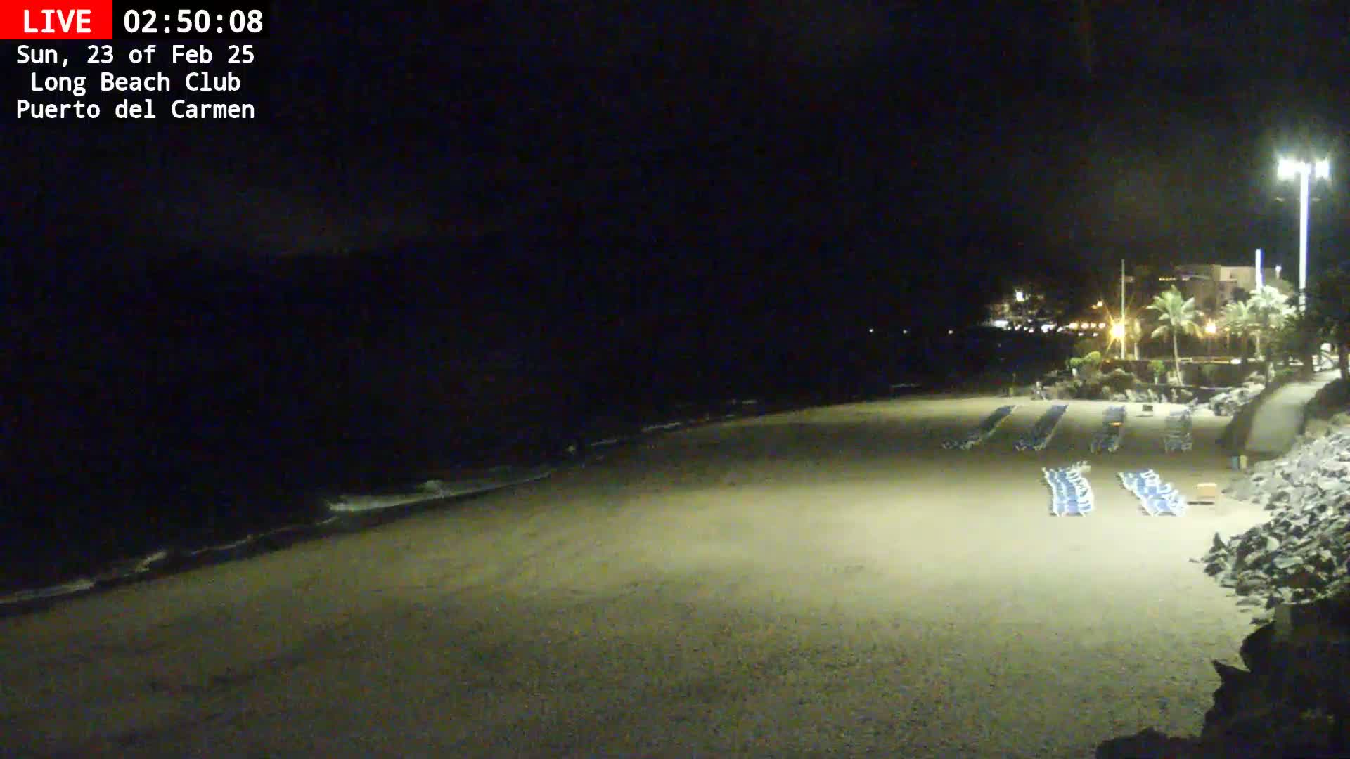 A mostly empty sandy beach at night is lit by artificial lights, with calm waves lapping the shore and rows of empty lounge chairs visible near a building in the distance.