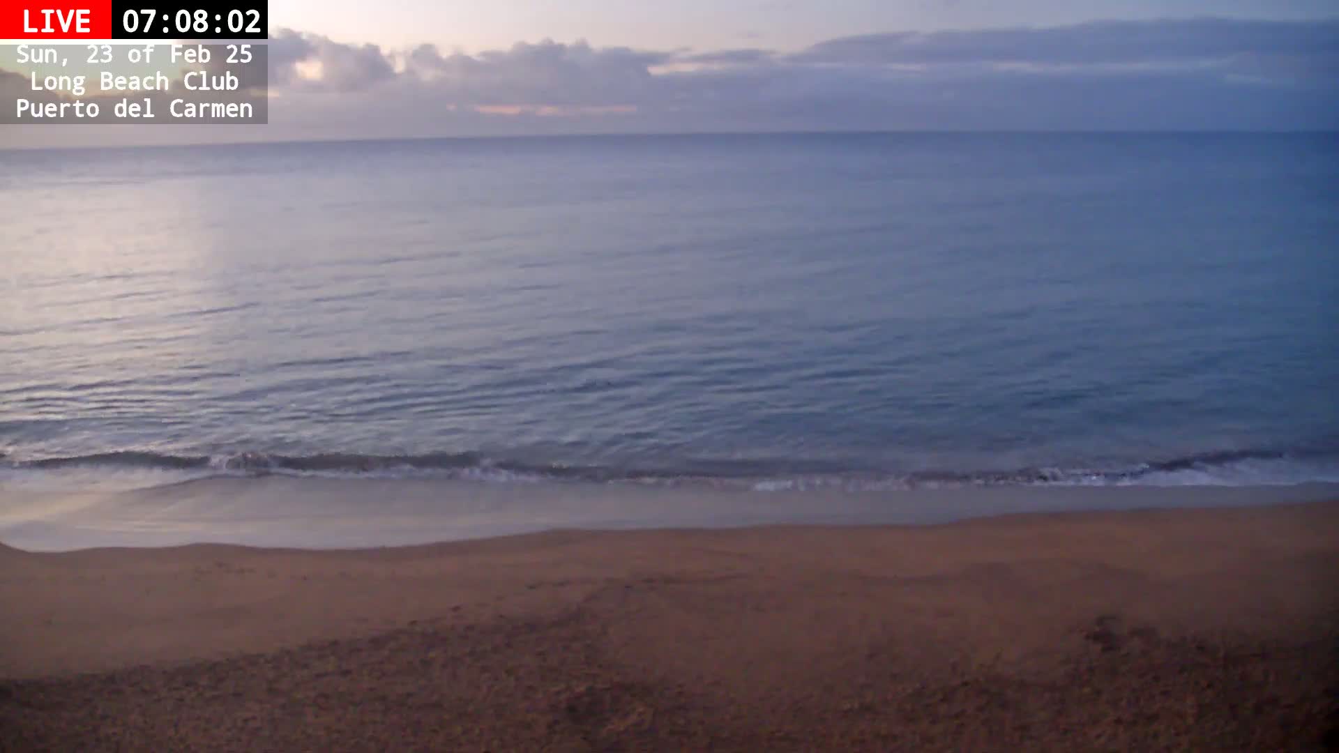A sandy beach meets a calm, blue ocean under a cloudy, dawn sky.