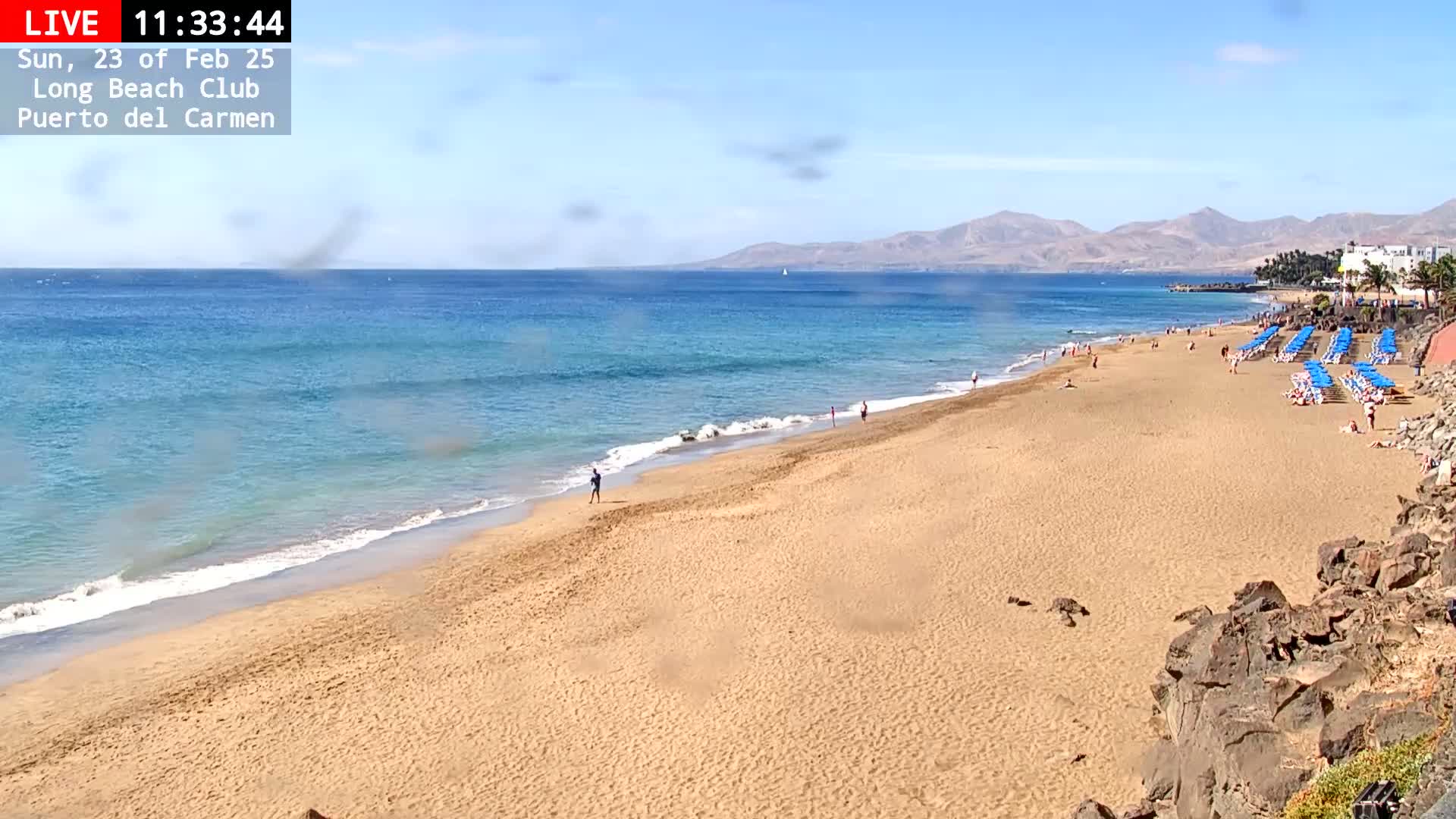 A sandy beach with light-blue water and some people stretches to mountains under a clear, sunny sky.