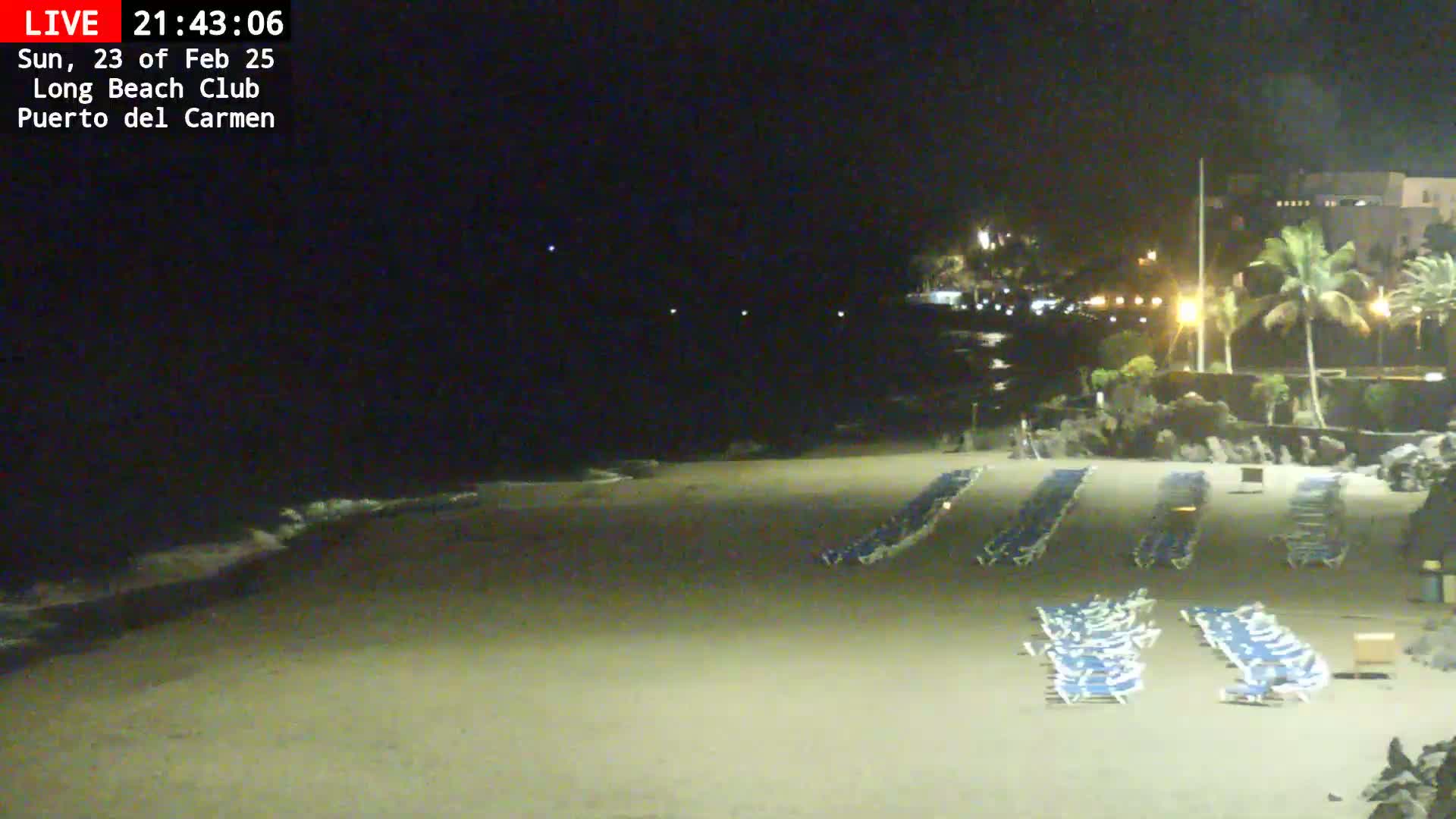 A dark sandy beach at night with rows of closed lounge chairs and a gently lapping ocean, lit by artificial lights from nearby buildings.