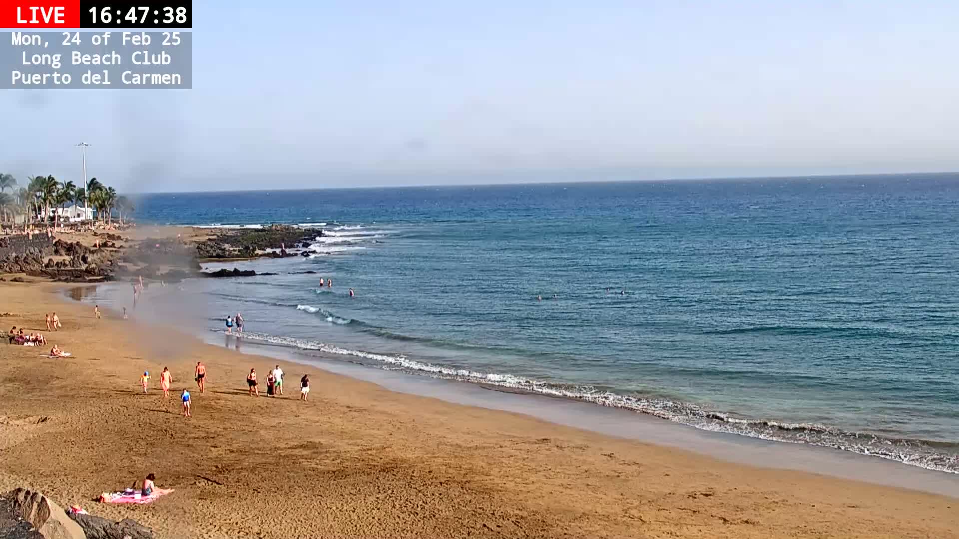 A sandy beach with a few people on it slopes gently down to a calm, blue ocean under a clear sky.