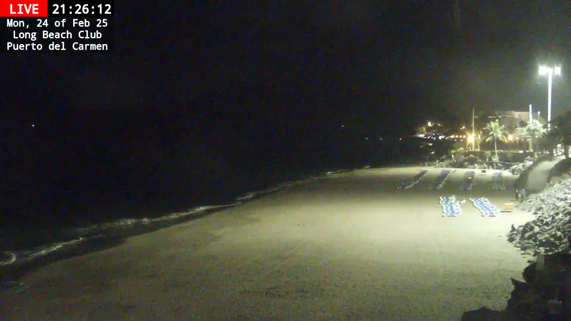 A dark, sandy beach at night with gentle waves lapping the shore, several rows of empty lounge chairs, and a few lights visible in the distance.